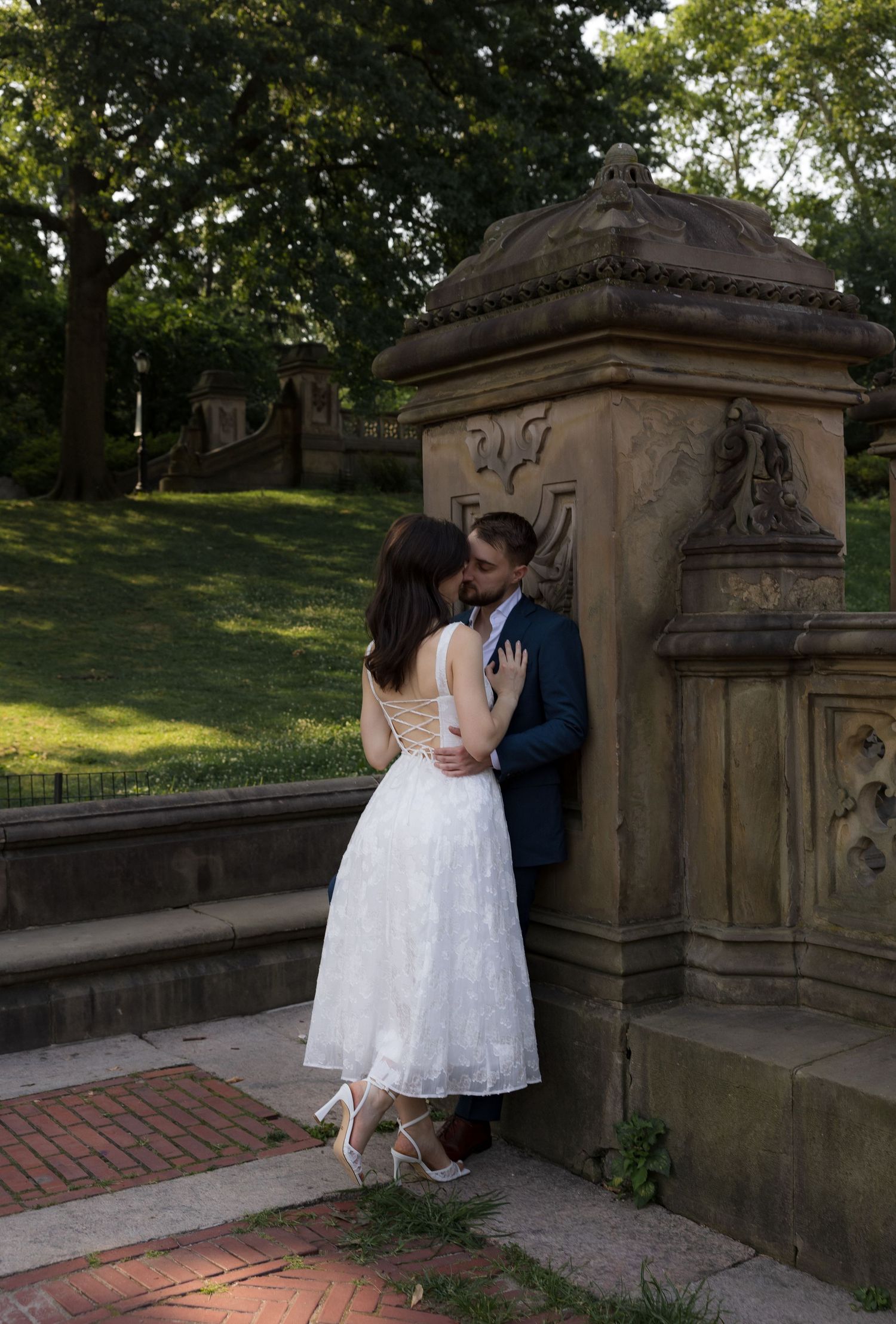 Couple sharing a romantic kiss in wedding attire beside an ornate stone pillar in a lush green park.