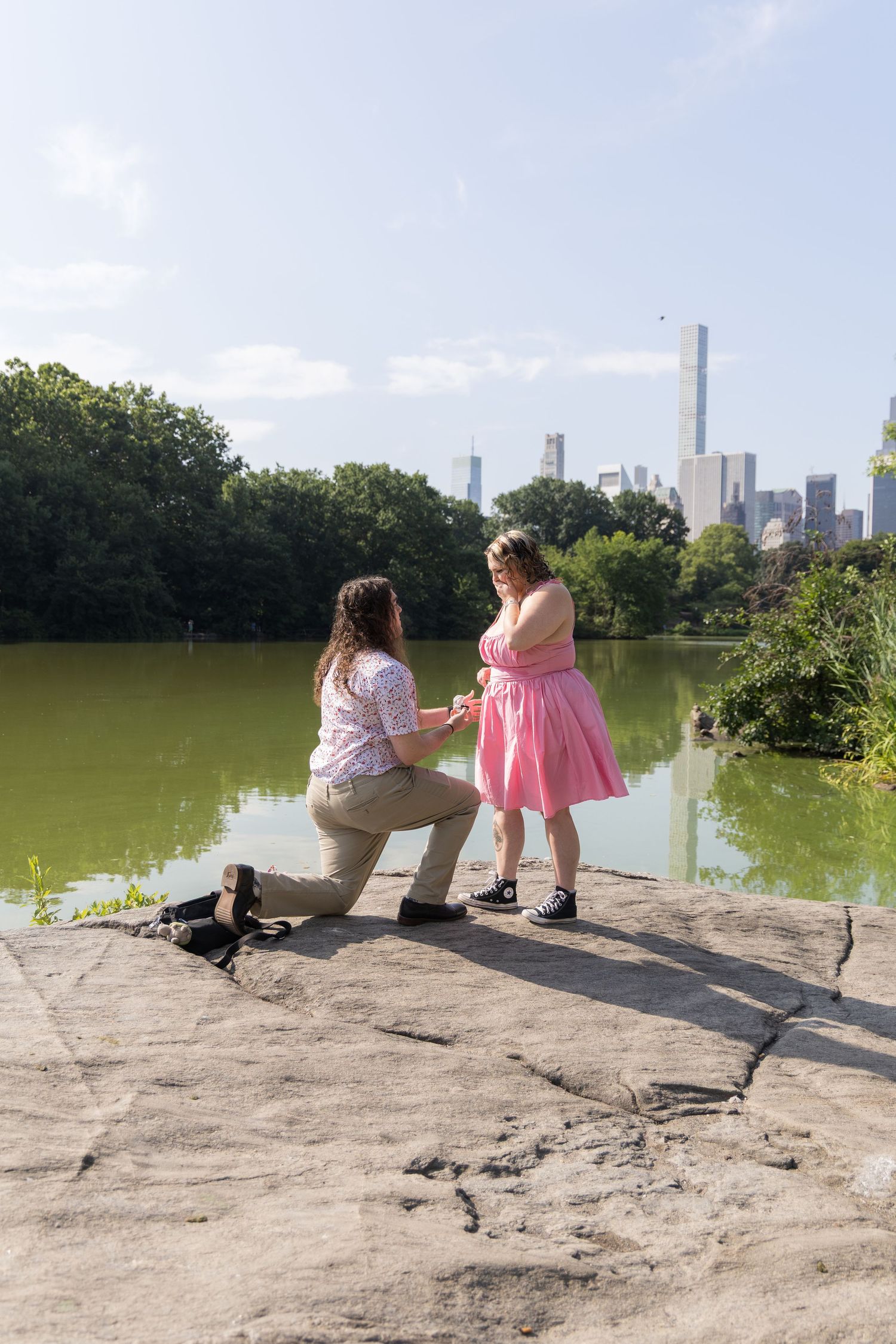 Same-sex proposal in Central Park NYC, one woman kneeling with ring as partner in pink dress looks on, skyline backdrop.