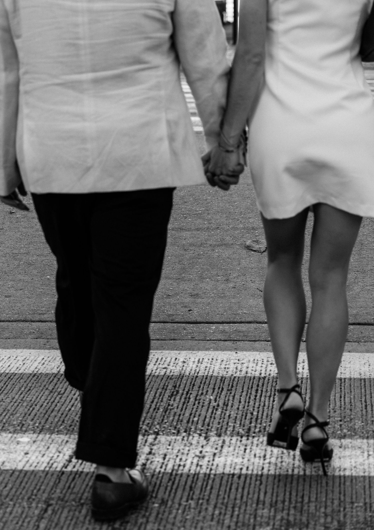 Black and white image of two people holding hands while walking, showing contrasting outfits of pants and a dress.