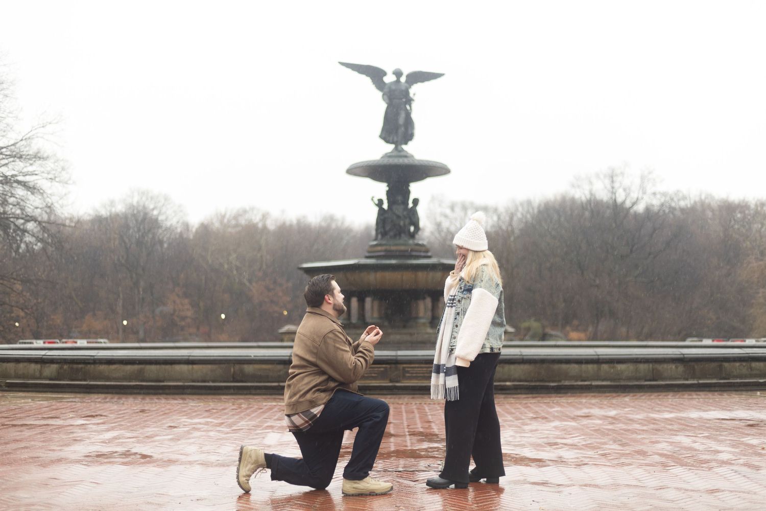 Man proposing to woman on one knee at Bethesda Fountain in Central Park on a rainy day.