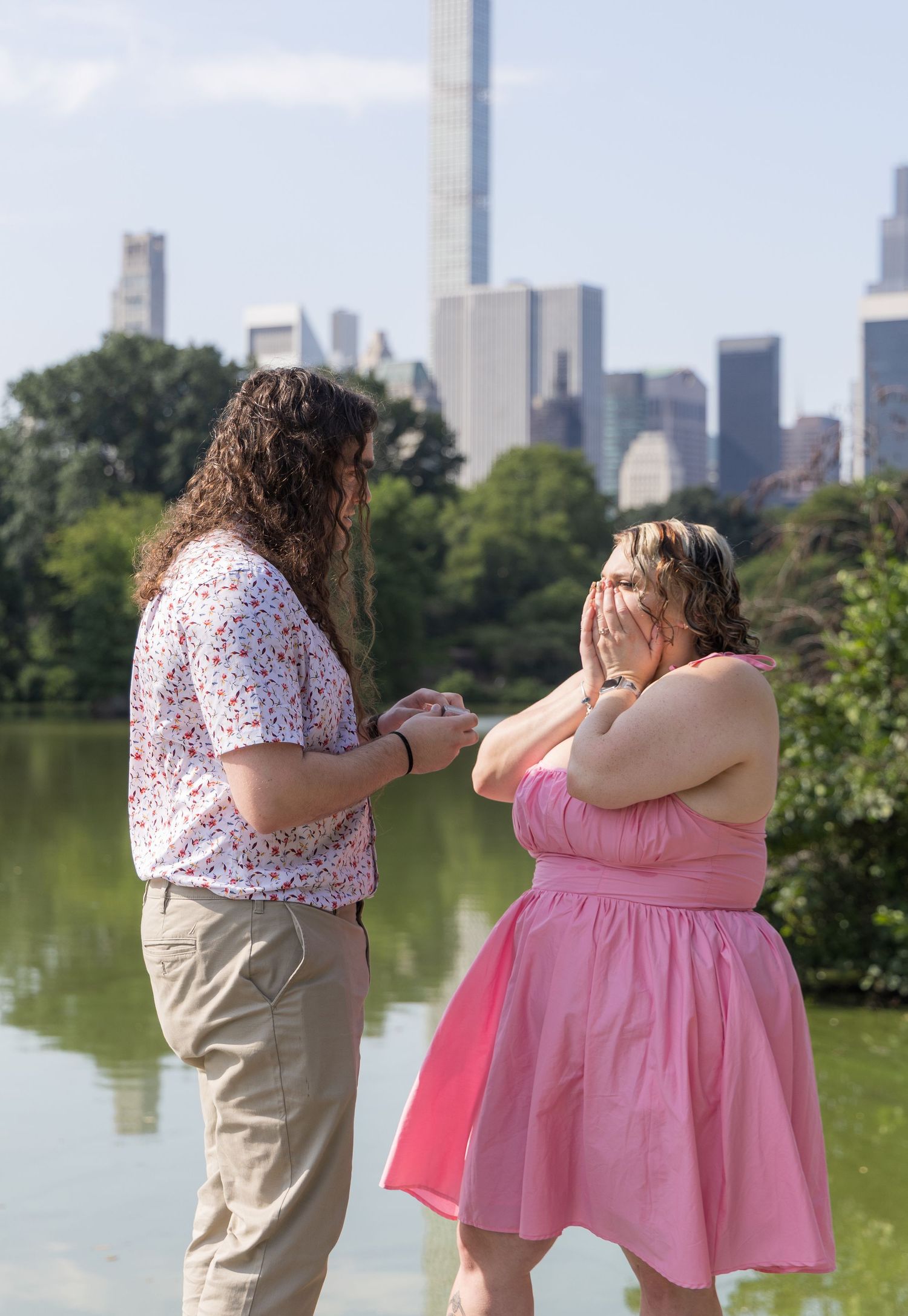A man proposes to a surprised woman in pink dress near a Central Park lake with NYC skyline backdrop.