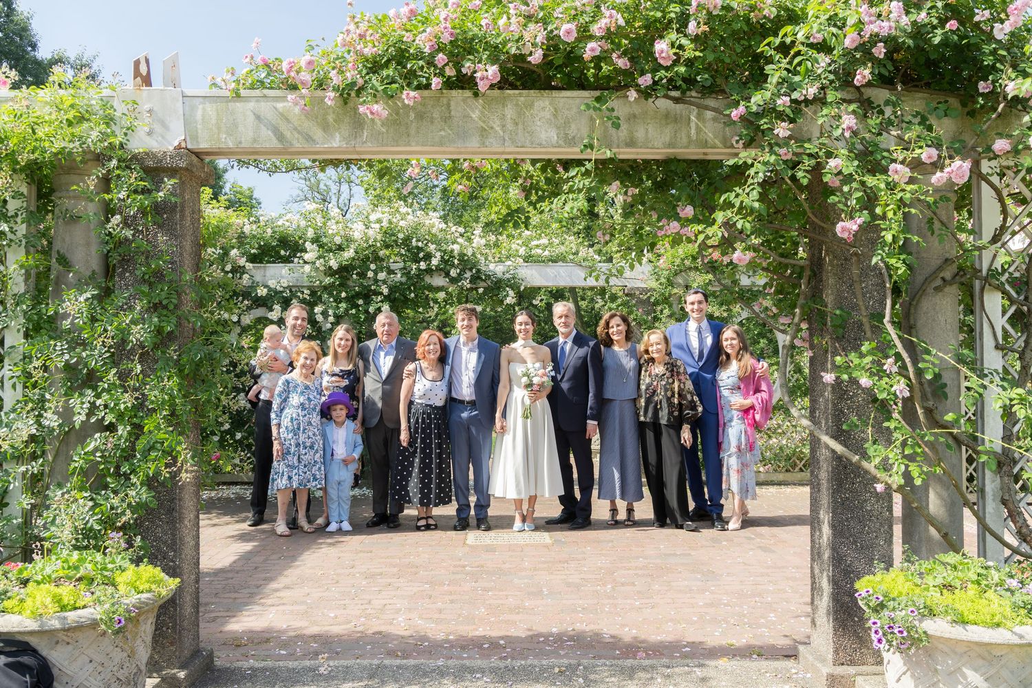 Group of elegantly dressed people posing under a flower-covered pergola in a lush garden setting.