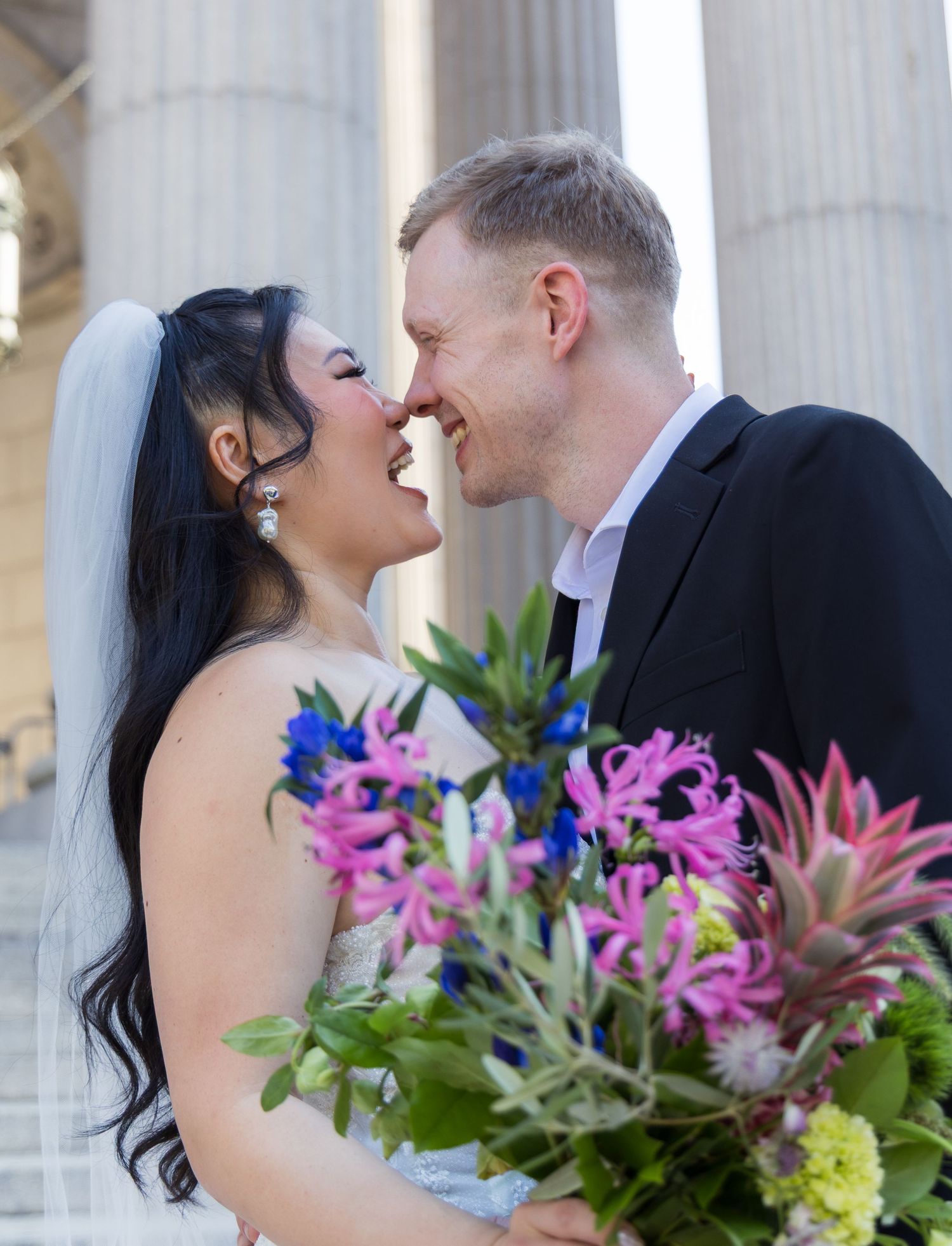 A couple shares an intimate moment among elegant columns with colorful floral arrangements in the foreground.