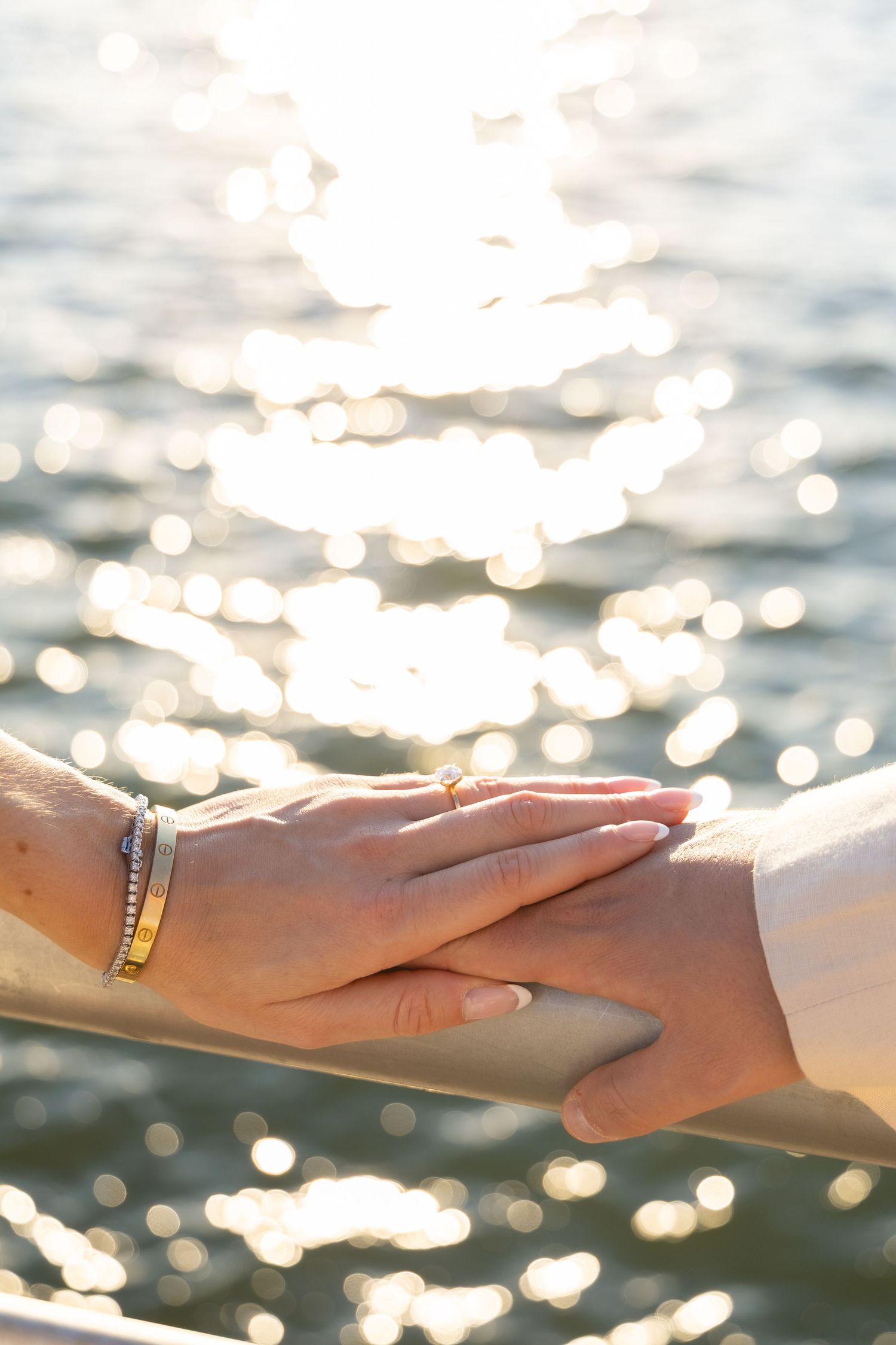 Two hands wearing wedding rings touch tenderly against a sparkling water background with bokeh light effects.