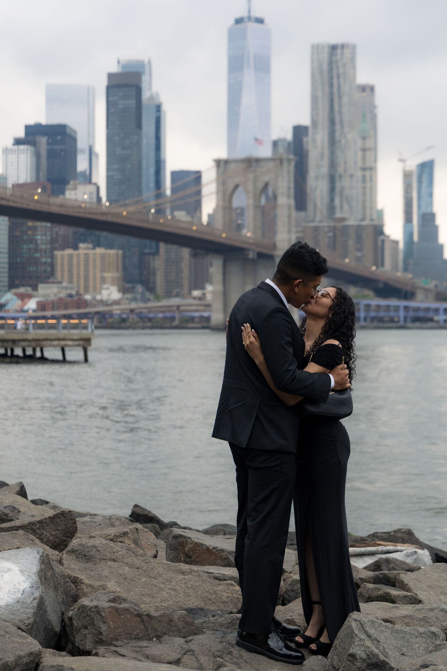 Couple embracing and kissing by the Brooklyn Bridge with NYC skyline backdrop, dressed in elegant black attire.