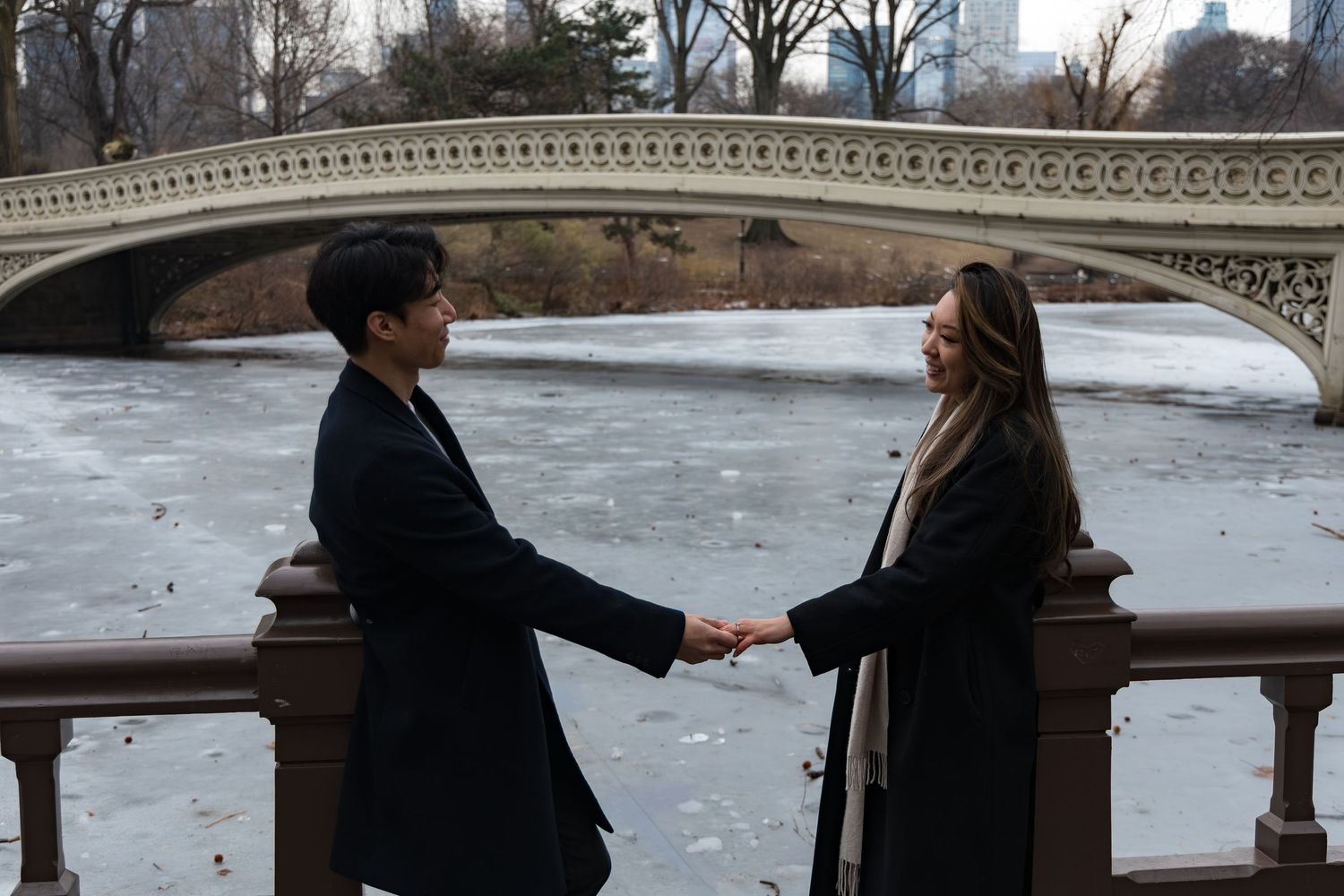 Couple holding hands by Bow Bridge in Central Park, NYC, wearing black coats with frozen lake backdrop.