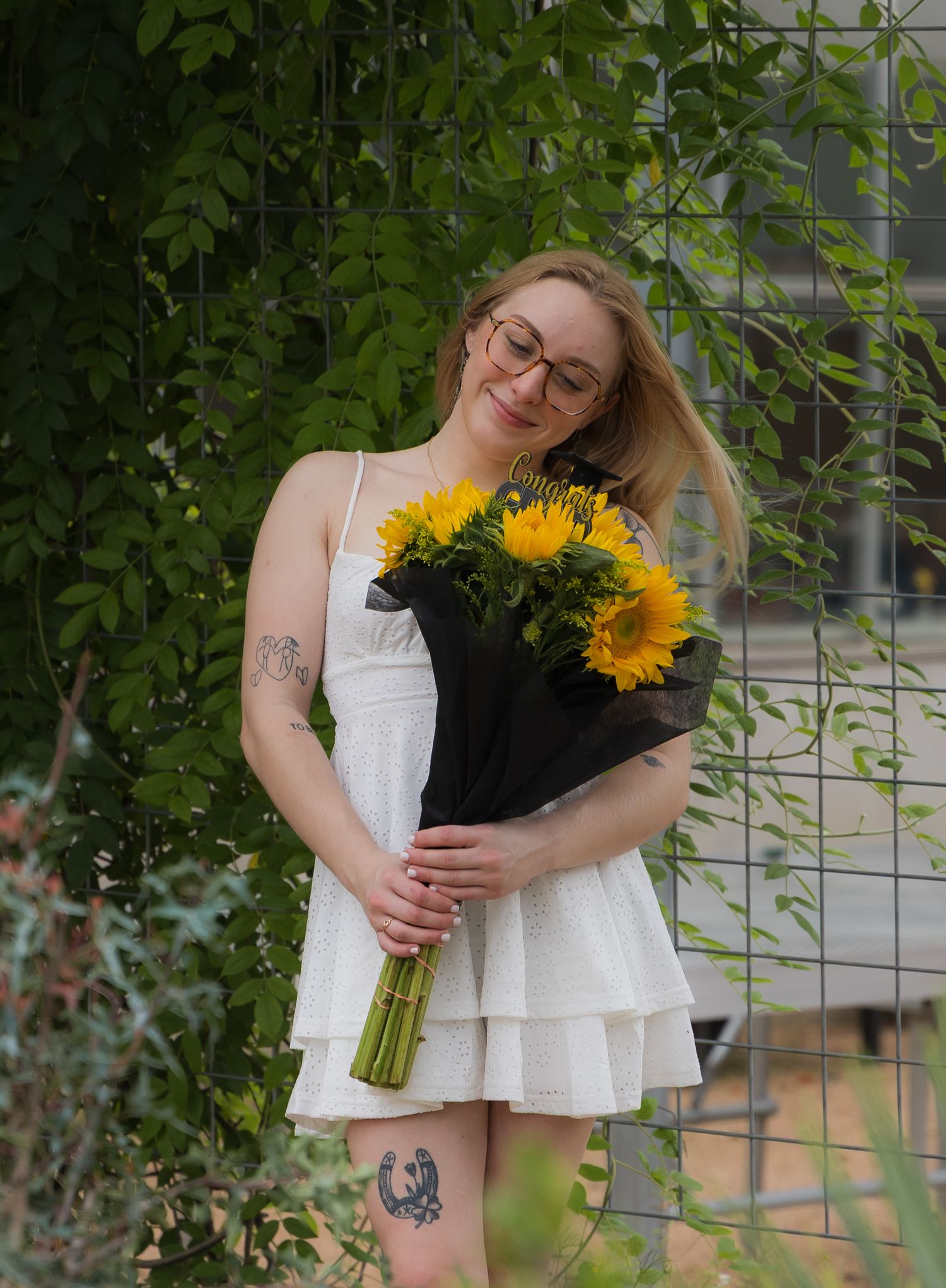 Smiling blonde woman in white dress holding sunflower bouquet beside green leafy fence.