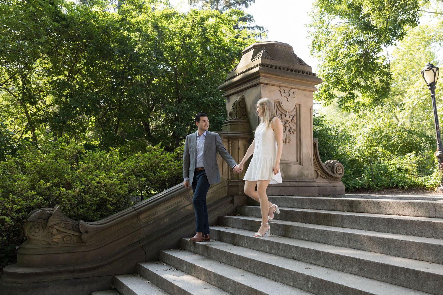 Couple holding hands walking down stone steps beside a decorative stone structure surrounded by lush green trees.