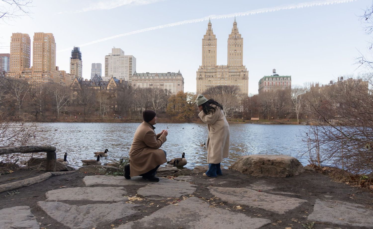 Two people sit on rocks by the lake in Central Park with the iconic San Remo towers and Manhattan skyline in winter.