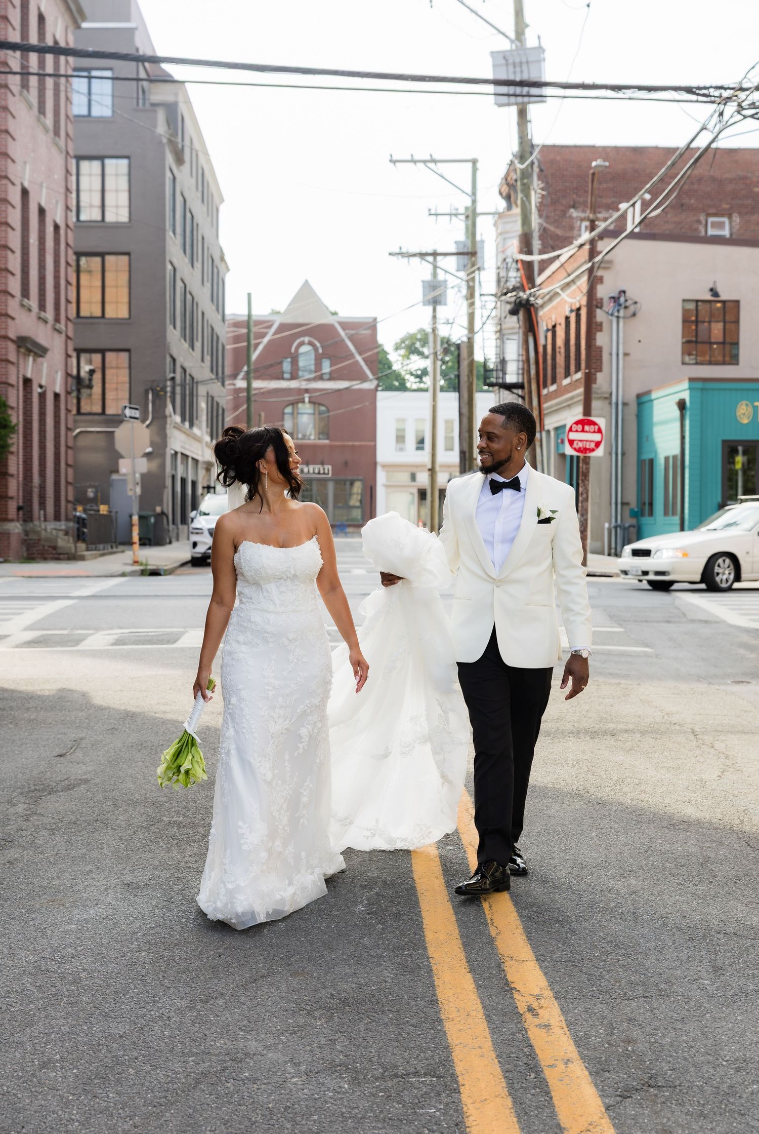 Bride and groom walking across urban street, she in strapless white gown, he in white tuxedo jacket with black bow tie.