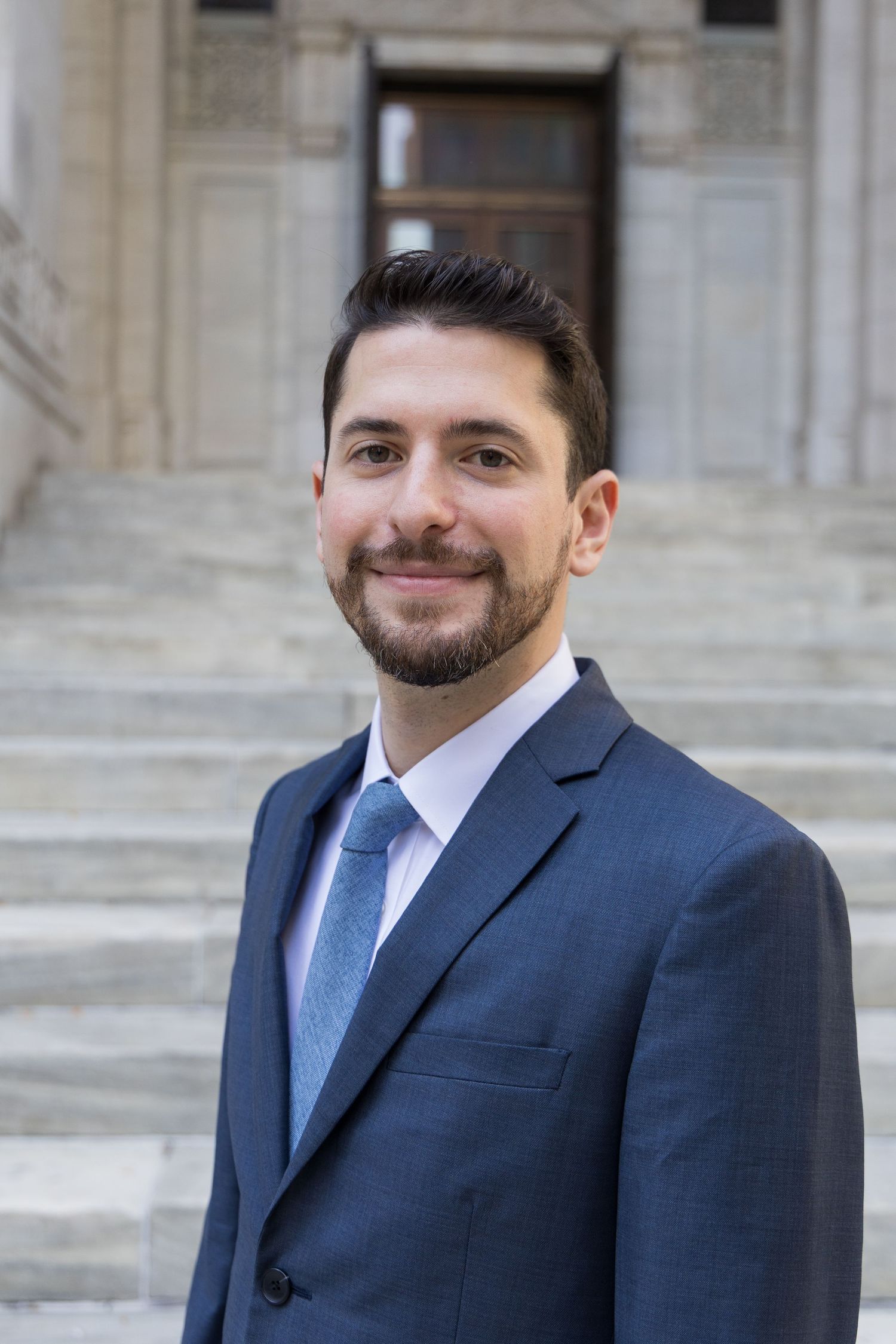 Professional headshot of someone in a navy suit and blue tie standing in front of a stone building exterior.
