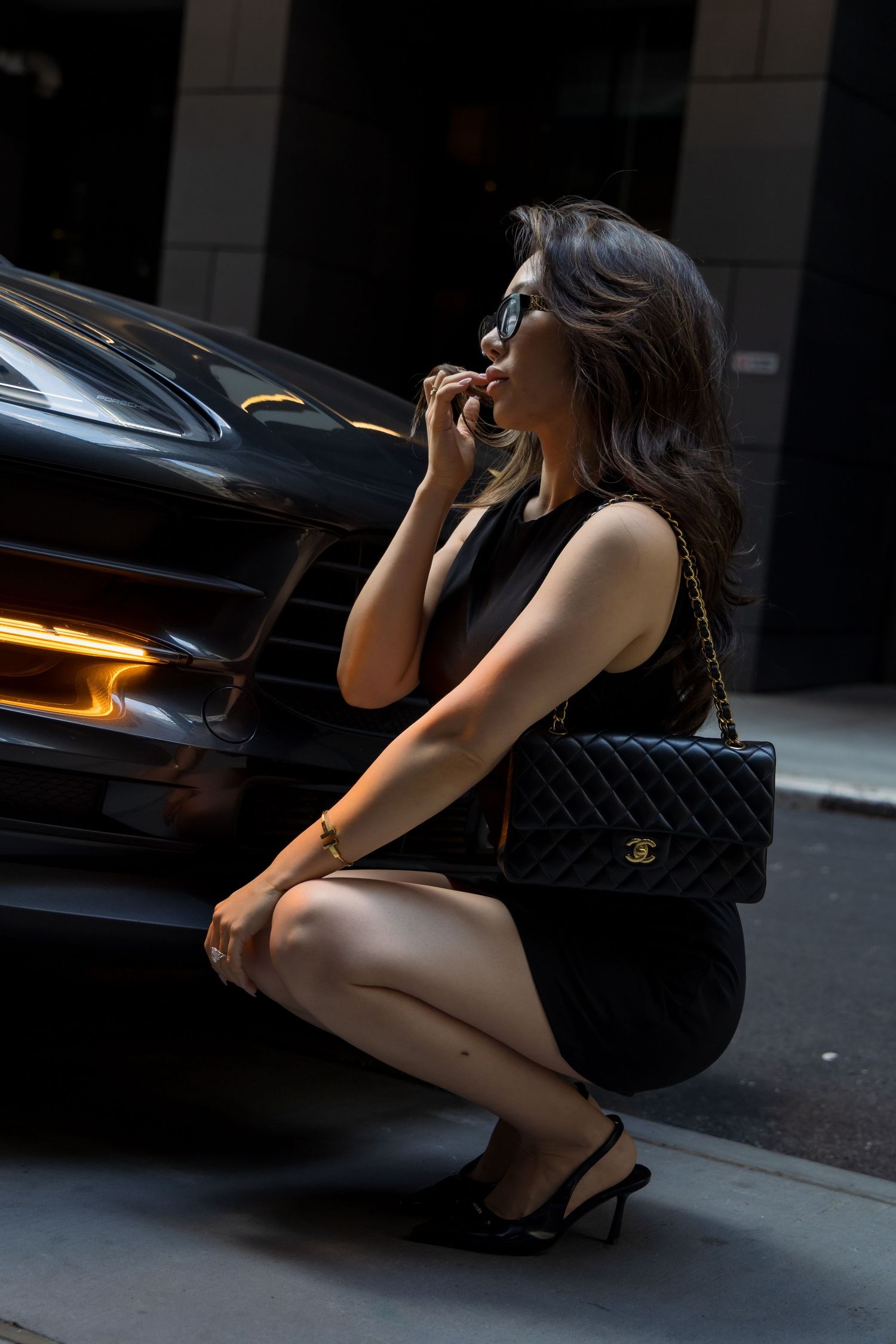 A woman in a black dress and heels crouches next to a car .