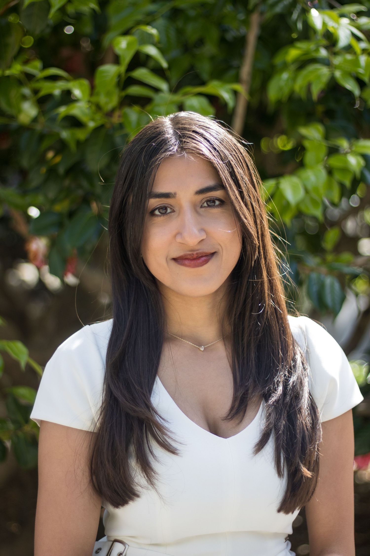 Woman with dark hair wearing a white top against a leafy background.