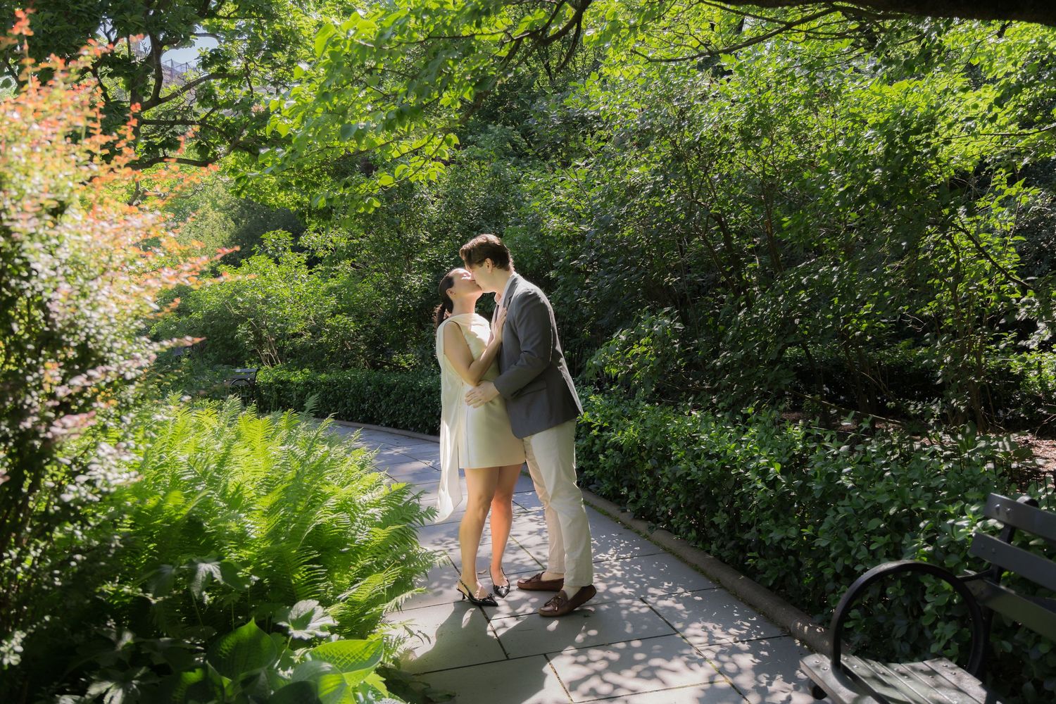 Couple sharing a romantic kiss on a garden path surrounded by lush green trees and foliage in a serene park setting.