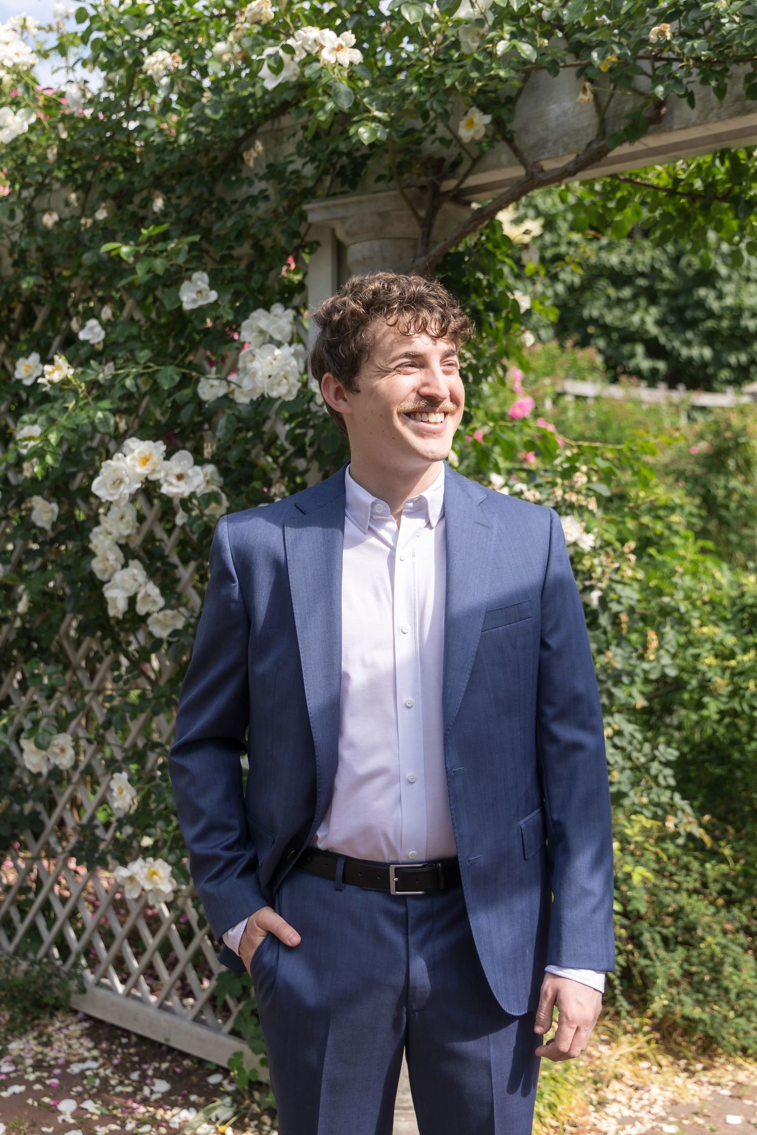 Young man in navy blue suit smiling outdoors near white flowering roses on a garden trellis.