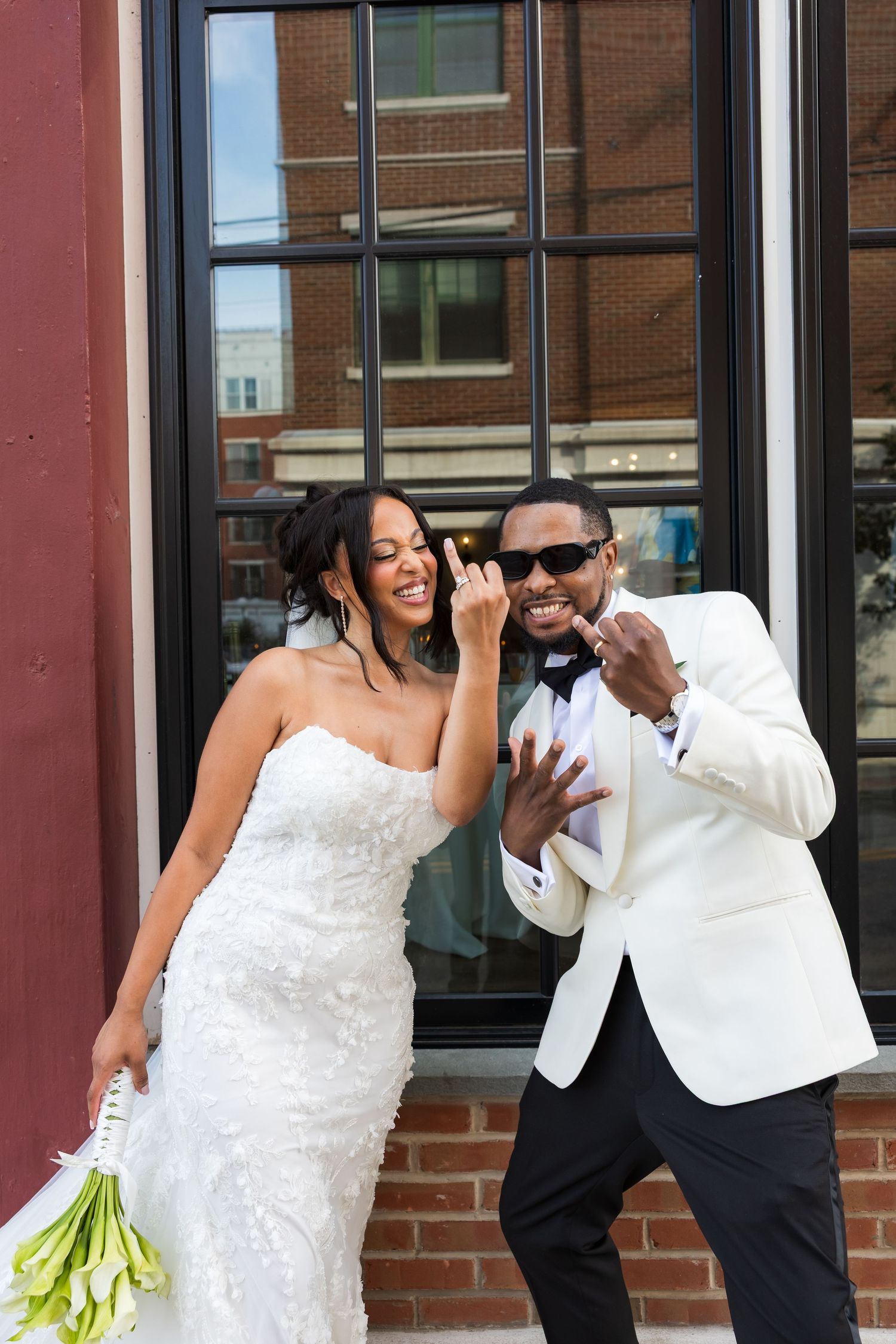 A joyful bride and groom in wedding attire pose playfully outside a brick building, showing off their wedding rings.