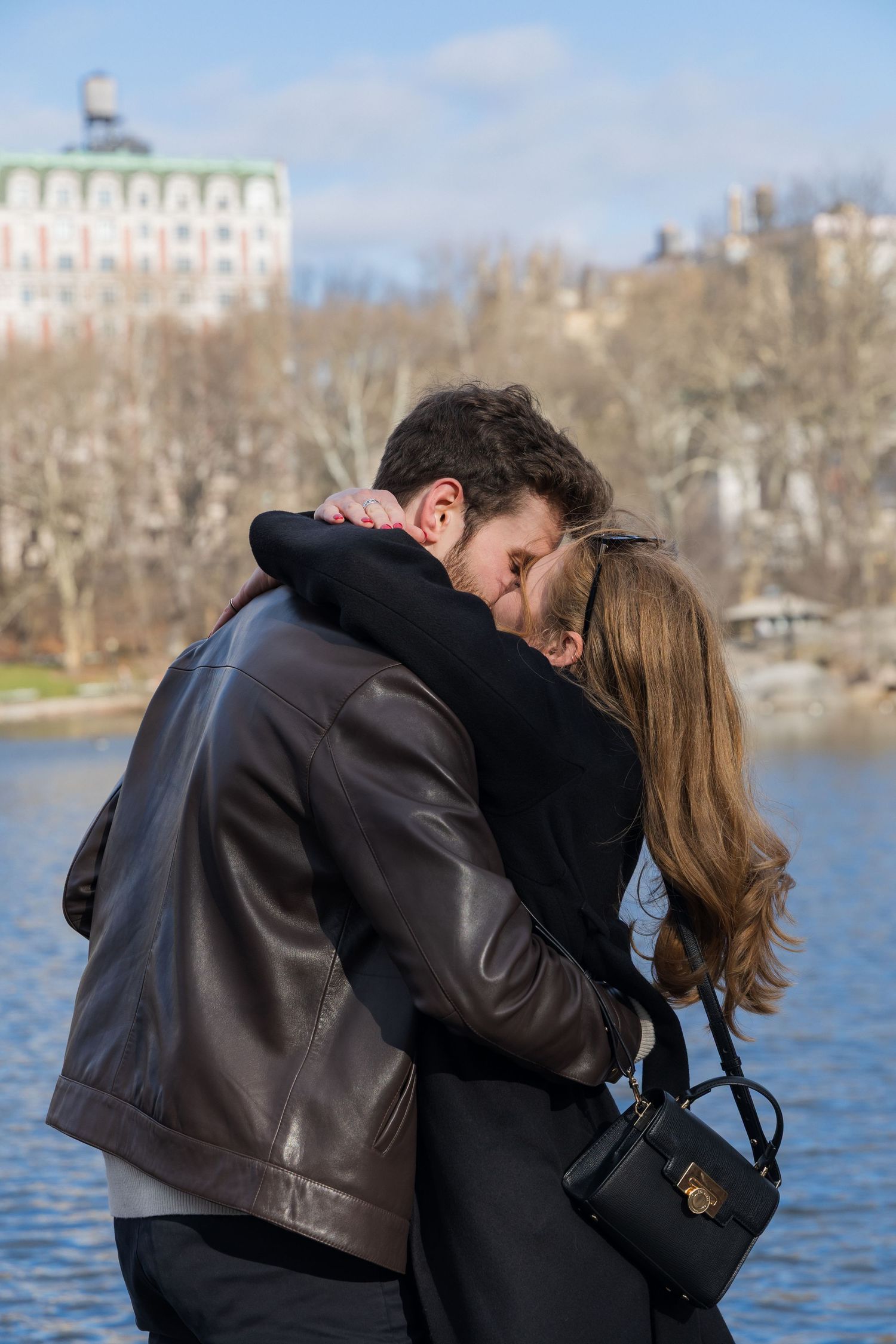 Couple sharing a romantic embrace and kiss near a lake in Central Park, New York, on a sunny winter day.