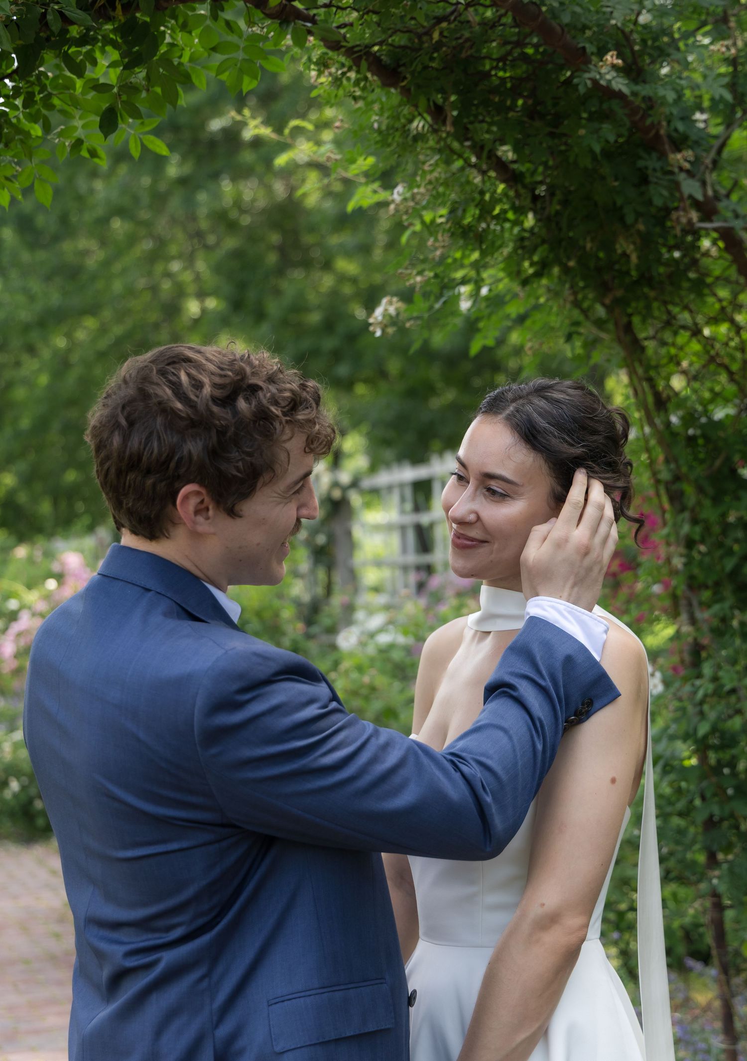 Groom in blue suit tenderly touches brides face in lush garden setting.
