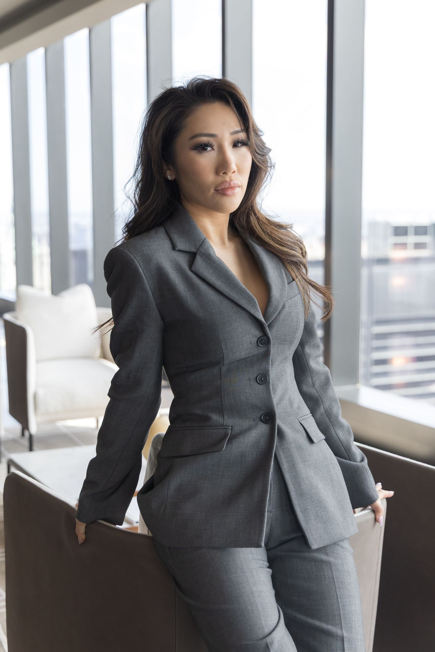 Professional woman in grey business suit poses in modern office space with large windows.