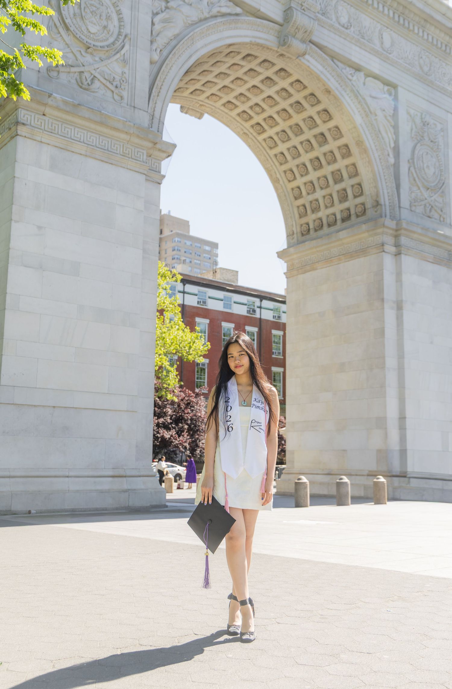 A graduate in white dress holds cap at Washington Square Park arch in NYC on a sunny day.