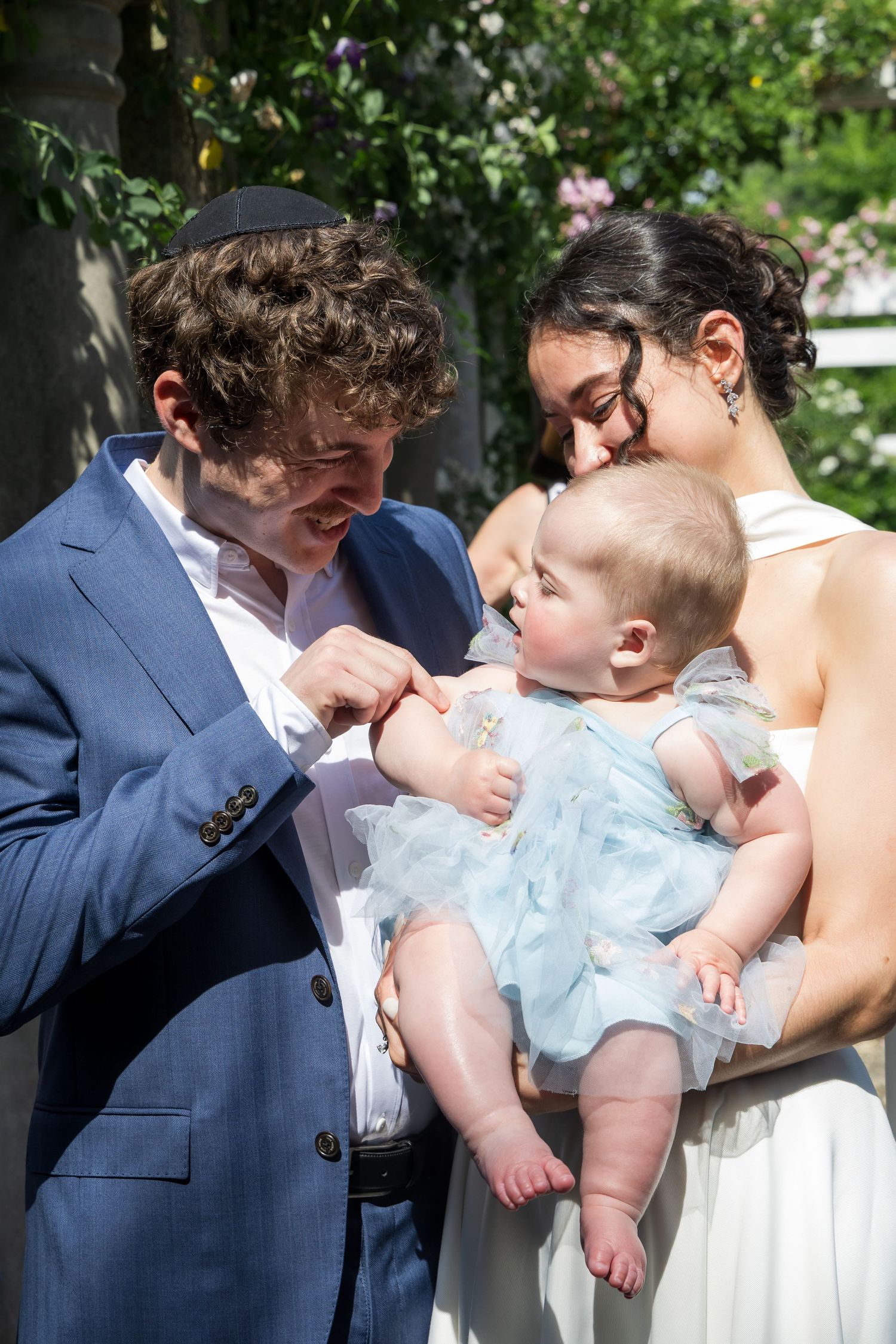A couple in formal attire tenderly interact with their baby dressed in a blue tulle outfit at an outdoor celebration.