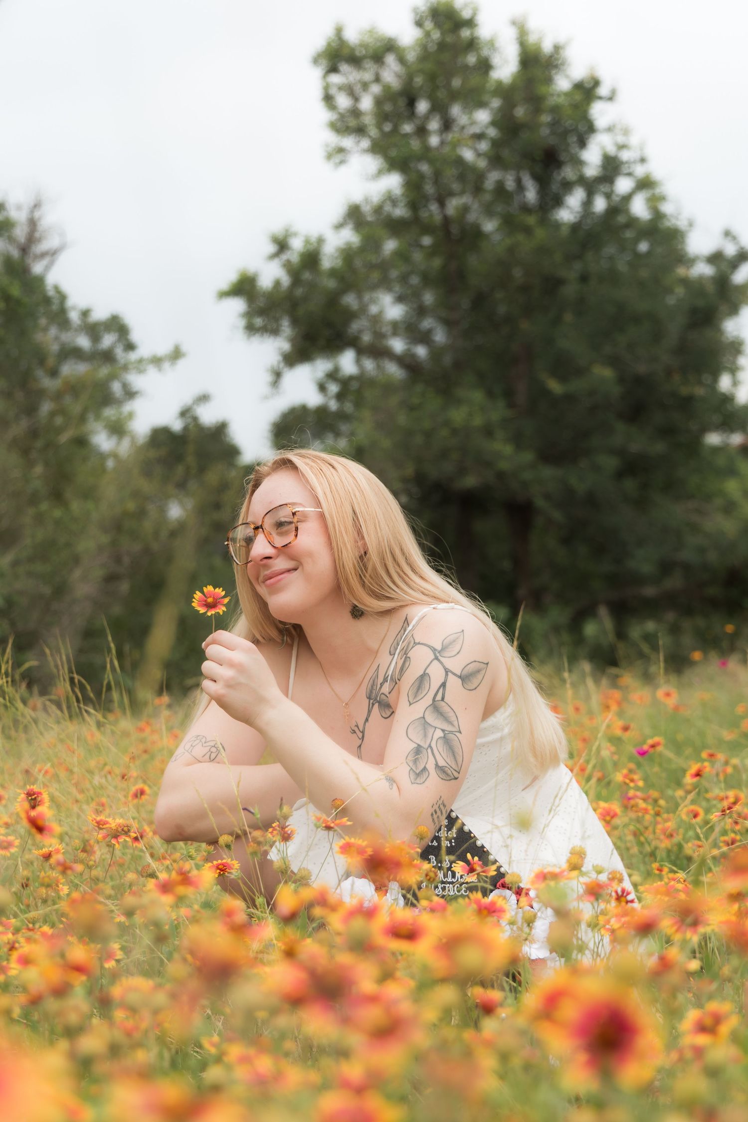 Smiling blonde woman with tattoos lying in orange wildflower field holding a small flower.