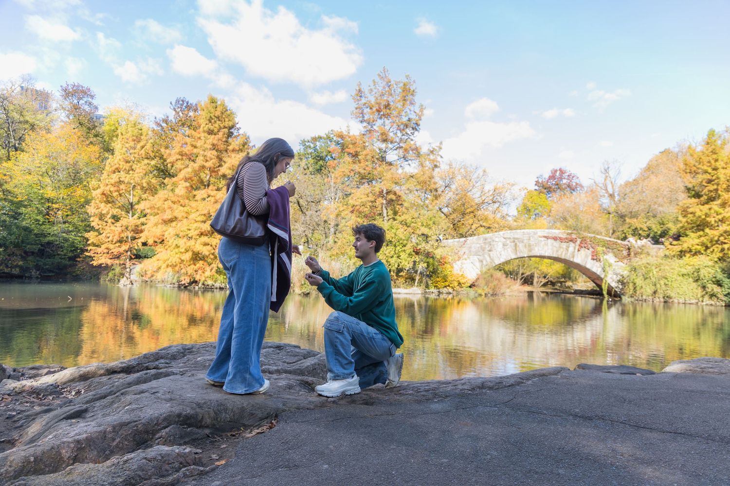 Man proposes to woman on one knee by a scenic autumn lake with colorful trees and stone bridge in Central Park.