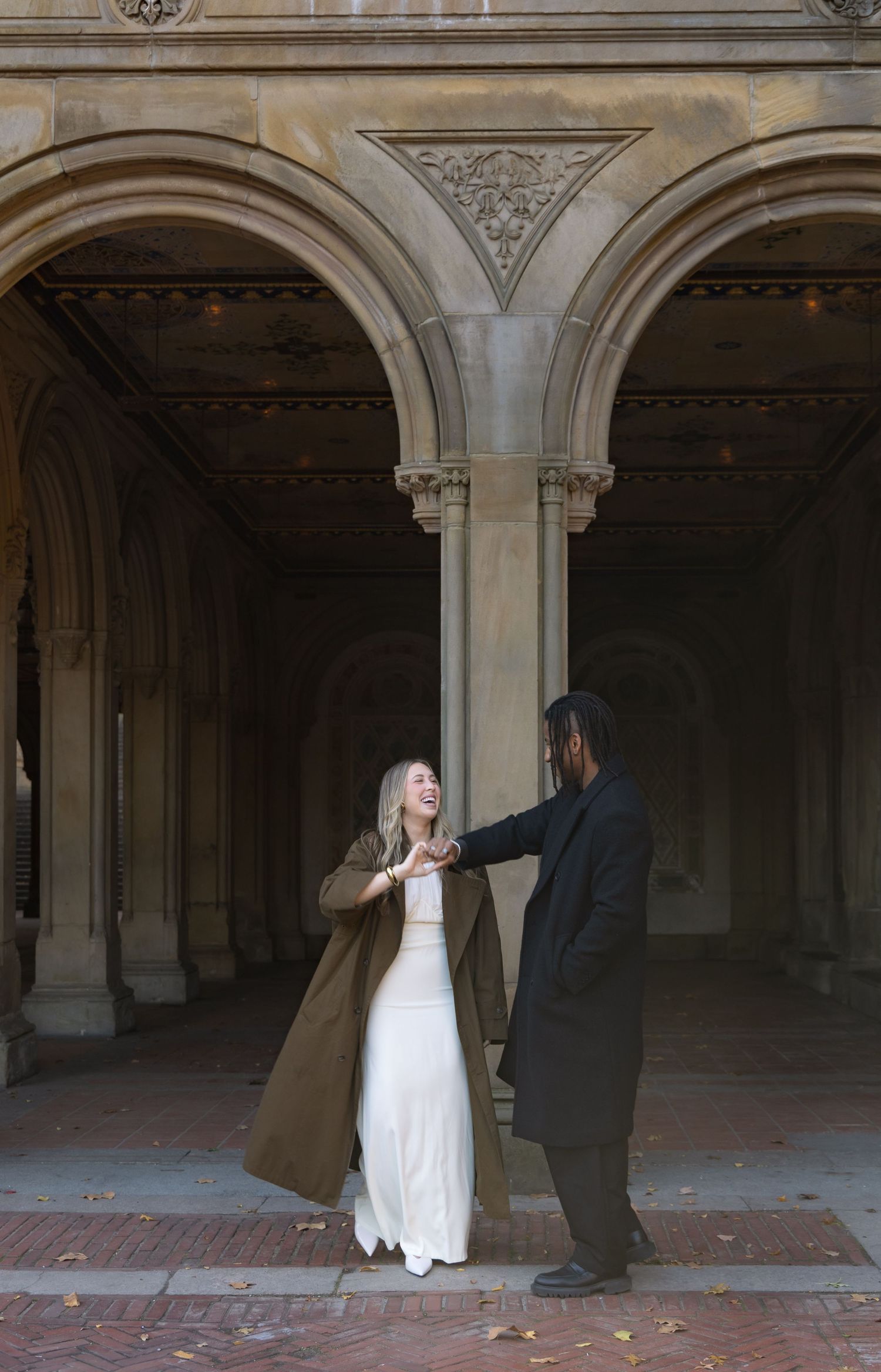 Couple dancing under stone arches at Bethesda Terrace in Central Park, bride in white gown and olive coat.