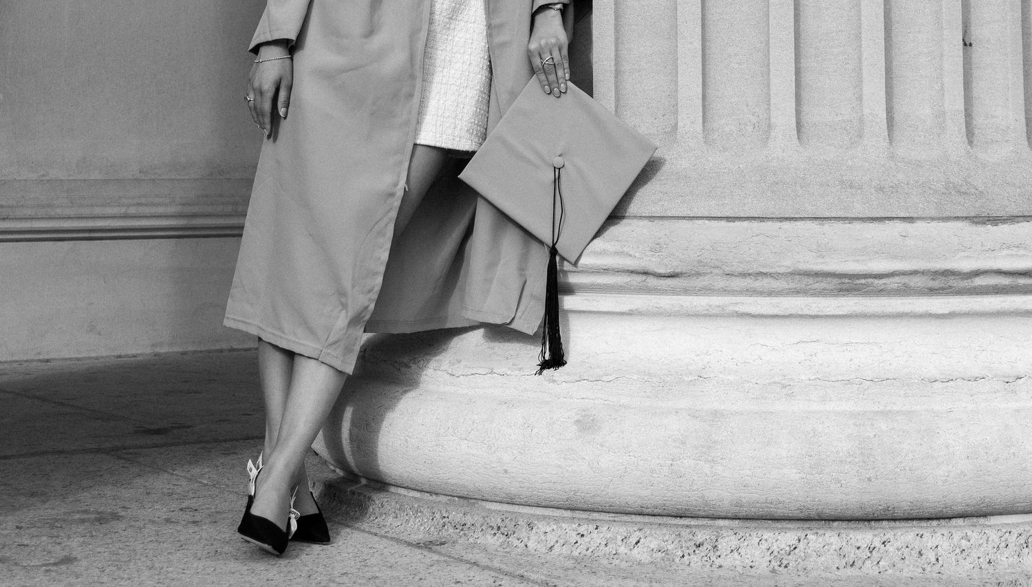 Black and white photo of a graduate holding a cap beside a stone column, wearing heels and a midi skirt.