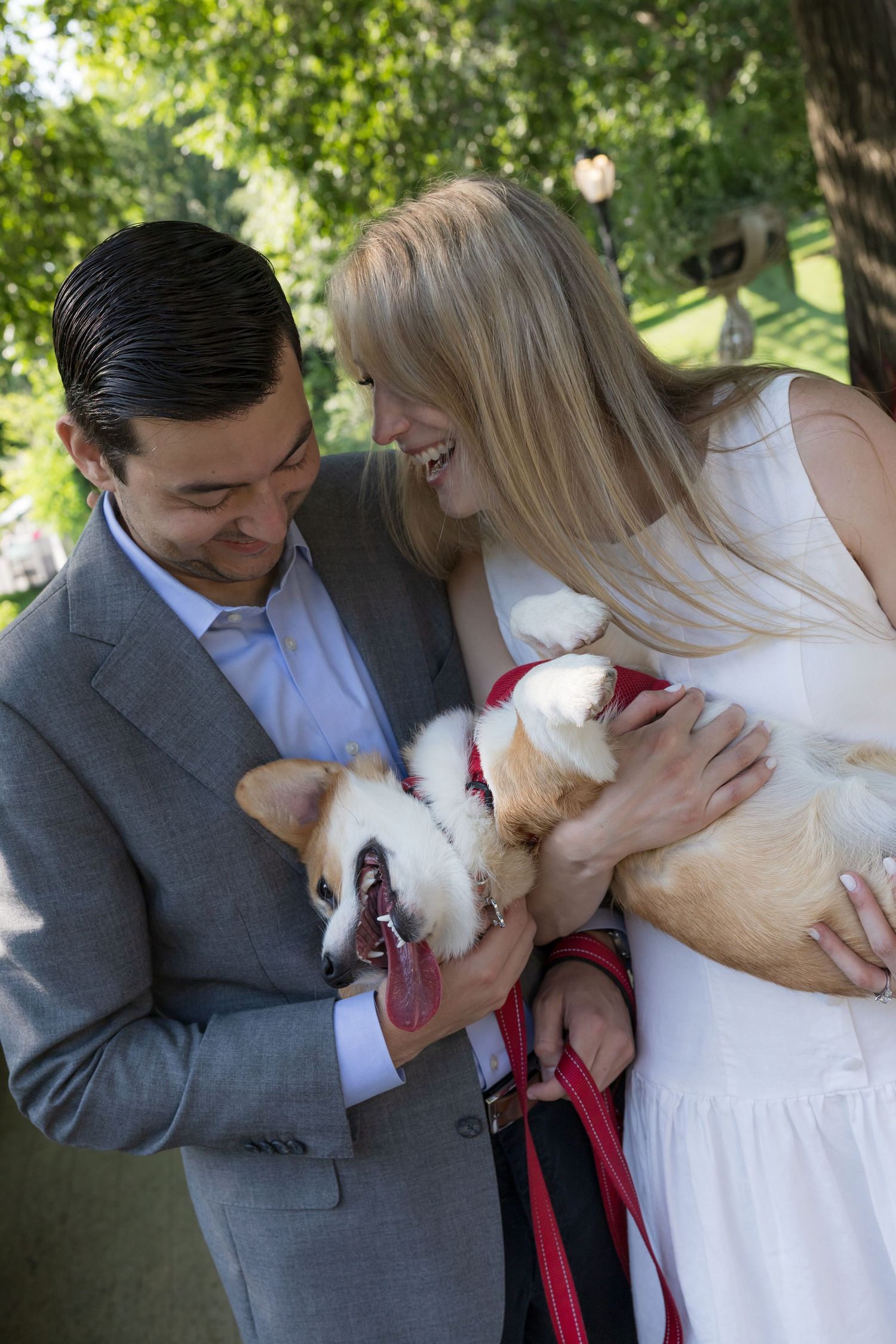 A happy couple laughs together while holding an adorable Corgi dog in a park.