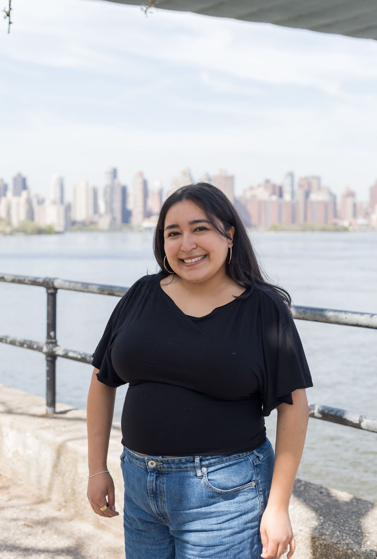 Smiling young woman in black top and jeans standing near waterfront railing with city skyline behind her.