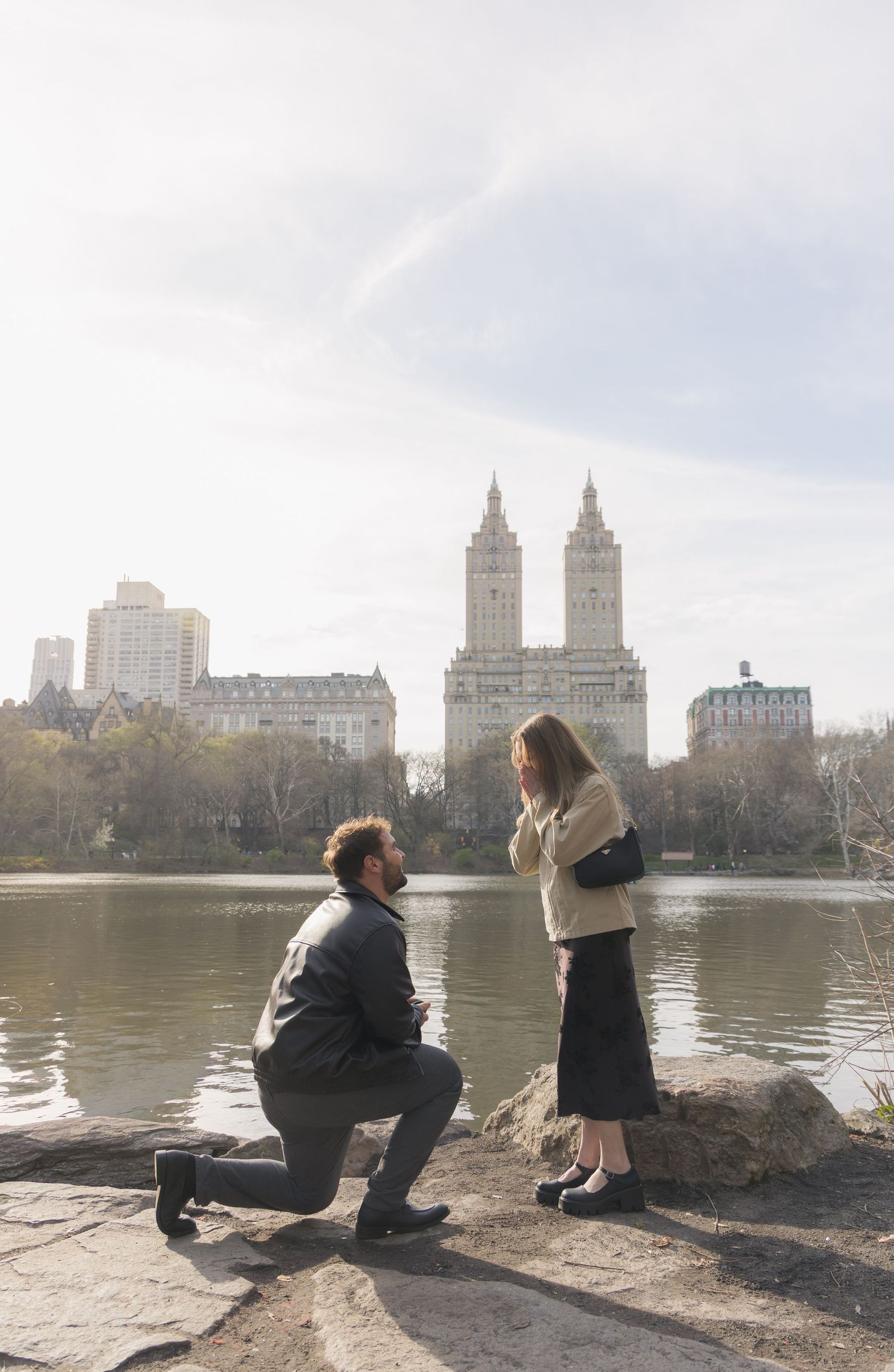 Man proposes on one knee to woman by Central Park lake with iconic San Remo building in NYC background.