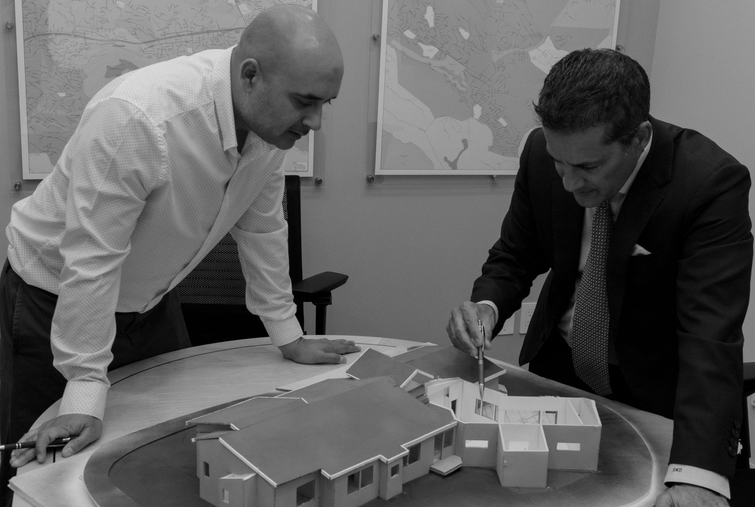 Two men in business attire examine architectural plans and a model layout on a table during a meeting.