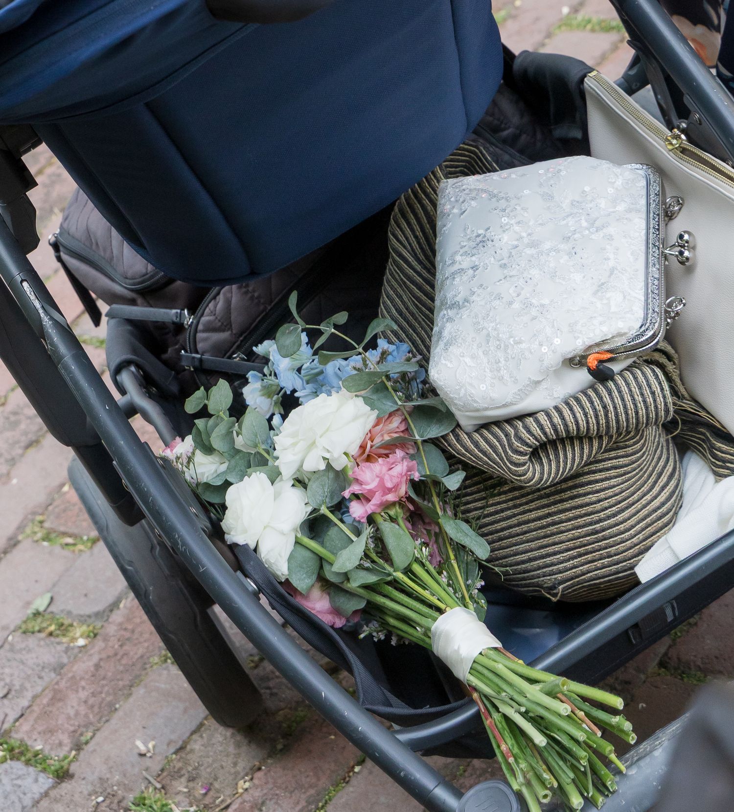 Baby stroller storage basket holding a bouquet of white and pink flowers, a striped bag, and a silver clutch purse.