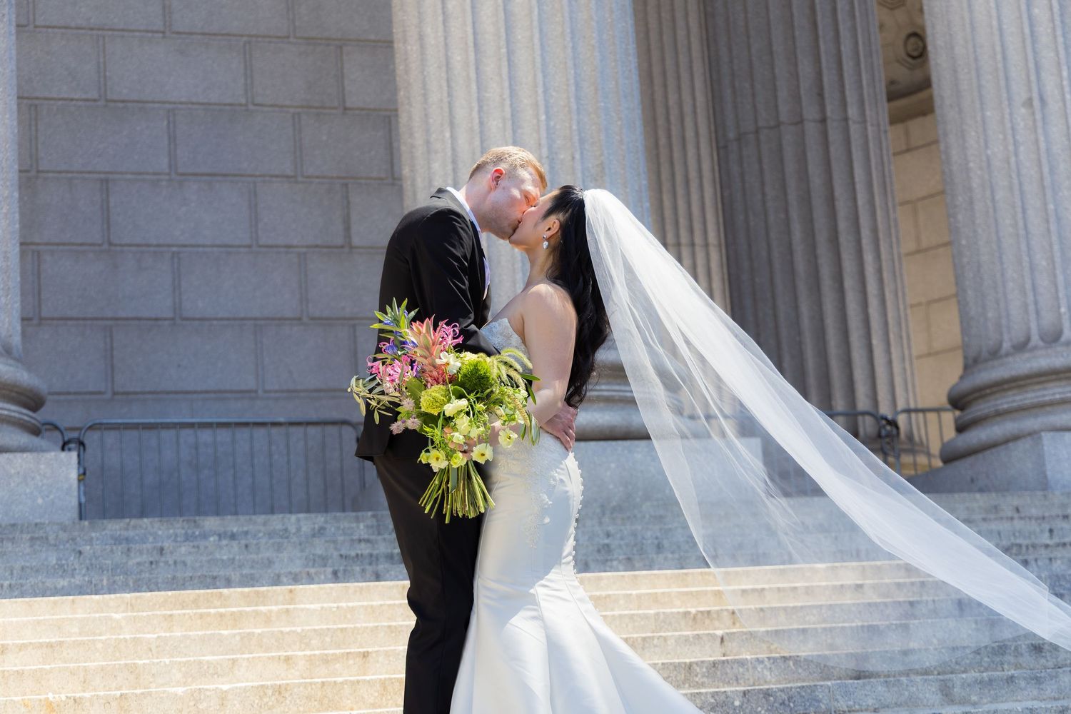 A couple shares a romantic kiss on stone steps of a grand building with white columns and brick details.