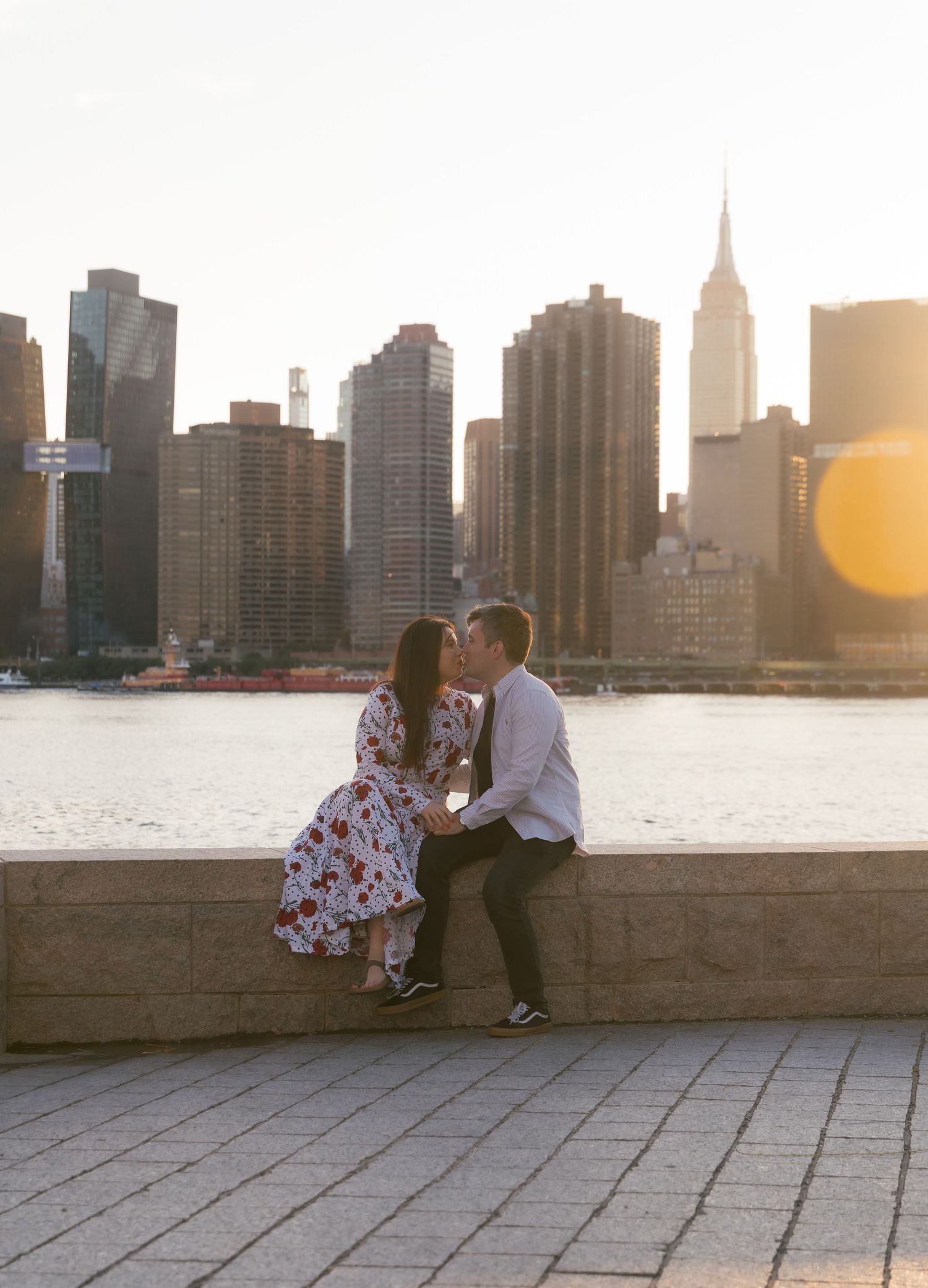 Couple sharing a romantic moment on NYC waterfront with Manhattan skyline and Empire State Building at golden hour.