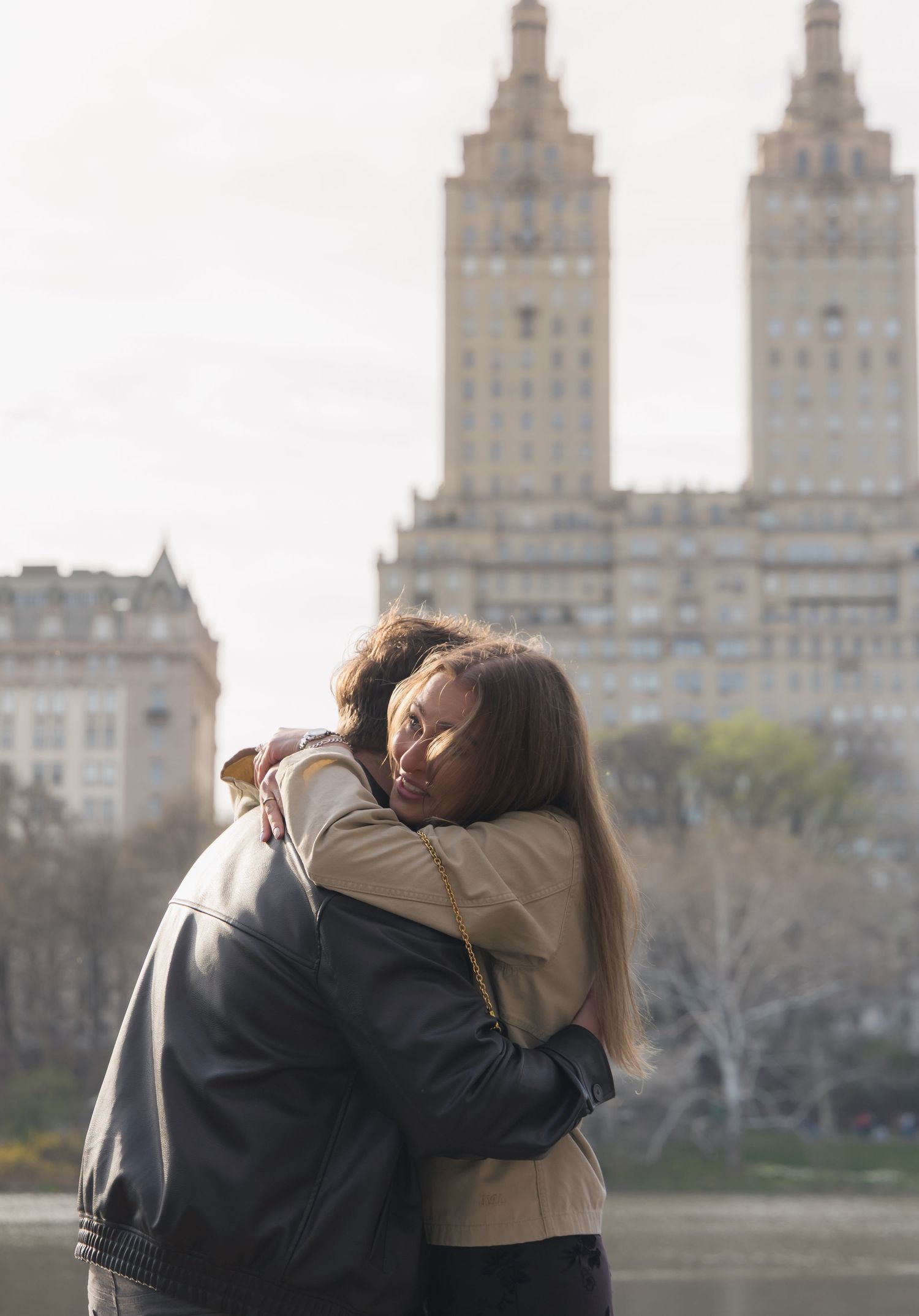 Two people sharing a warm embrace in Central Park with the iconic San Remo building in the background.