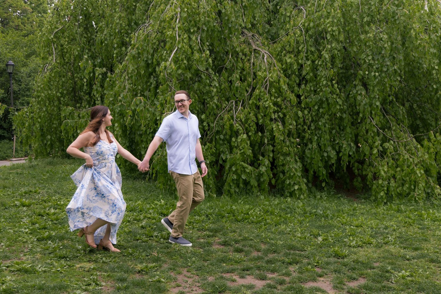 A couple holds hands and walks through a lush green park near a weeping willow tree on a sunny day.