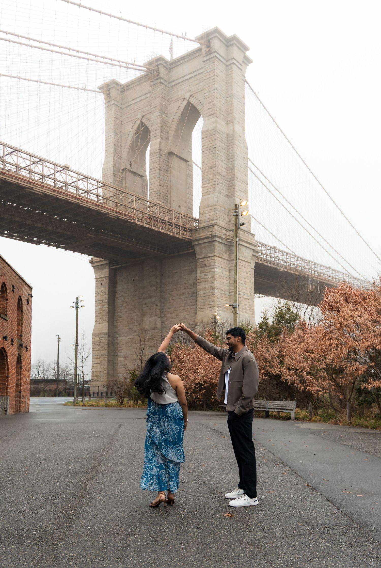 Couple dancing near Brooklyn Bridge in NYC, woman in blue skirt, man in gray jacket, cherry blossoms in background.
