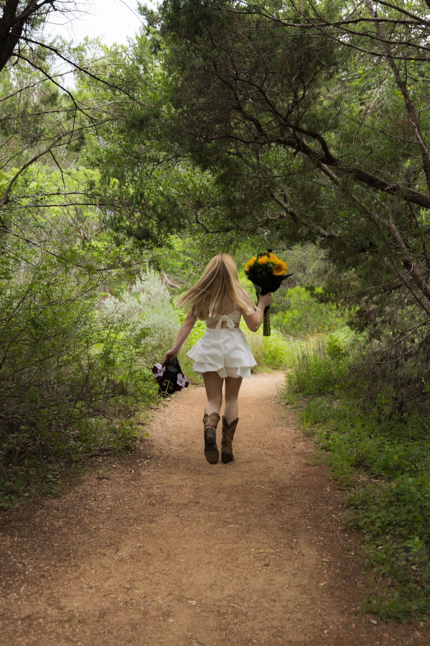 Blonde woman in white dress and cowboy boots walking down a wooded path holding flowers.