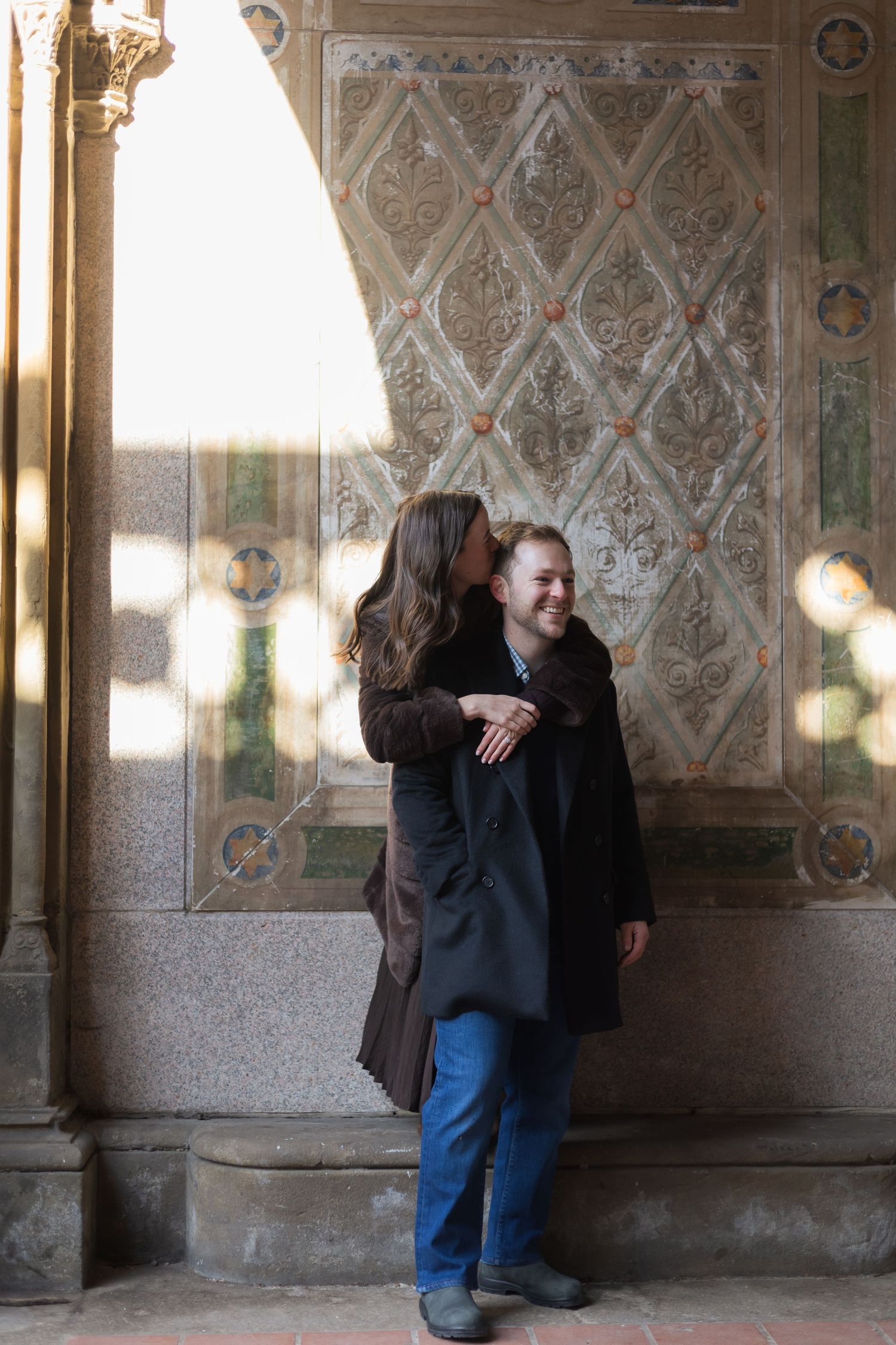 Two people stand together under an archway of Bethesda Terrace hugging.