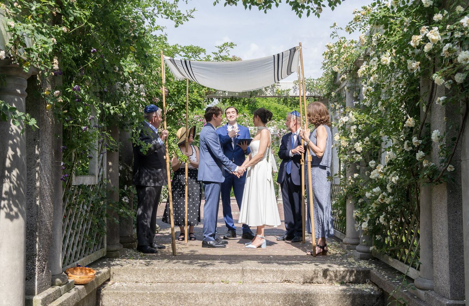 Couple exchanges vows under a floral chuppah surrounded by guests in a lush garden wedding ceremony.