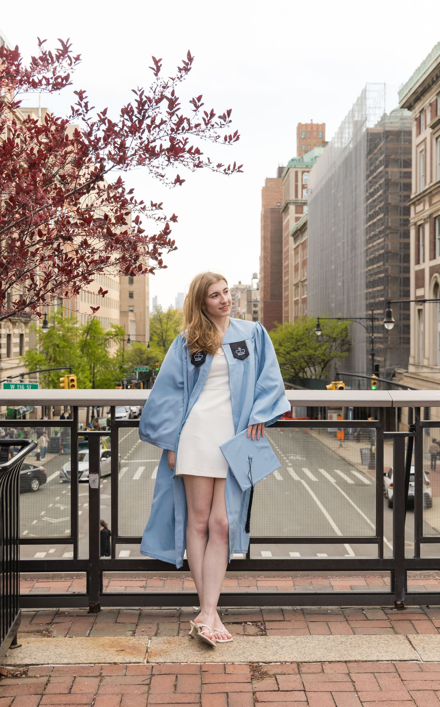 Young woman in graduation gown and white dress posing on urban balcony with city street and blooming tree backdrop.