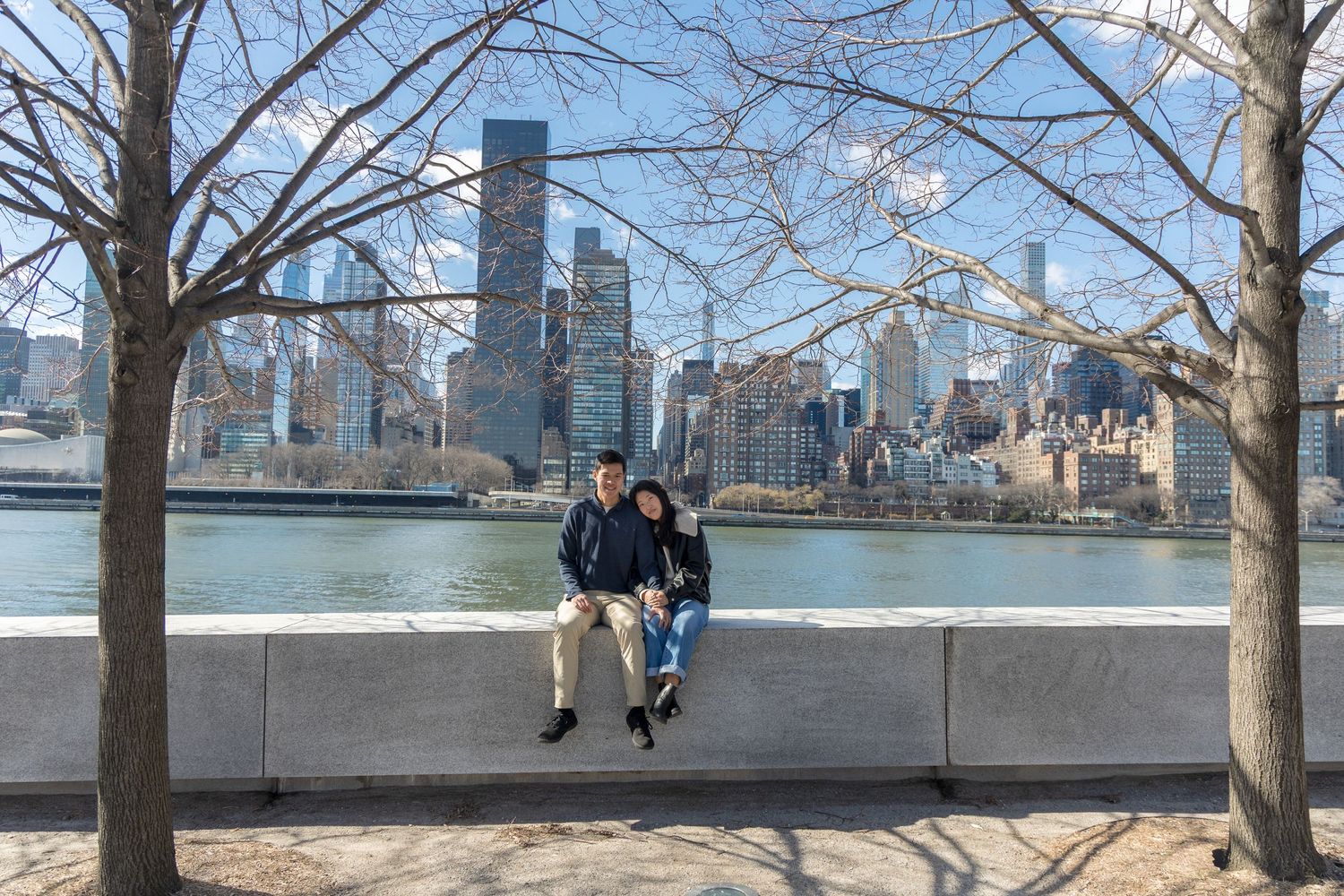 Couple sitting on a stone wall with NYC skyline and river backdrop on a clear winter day.