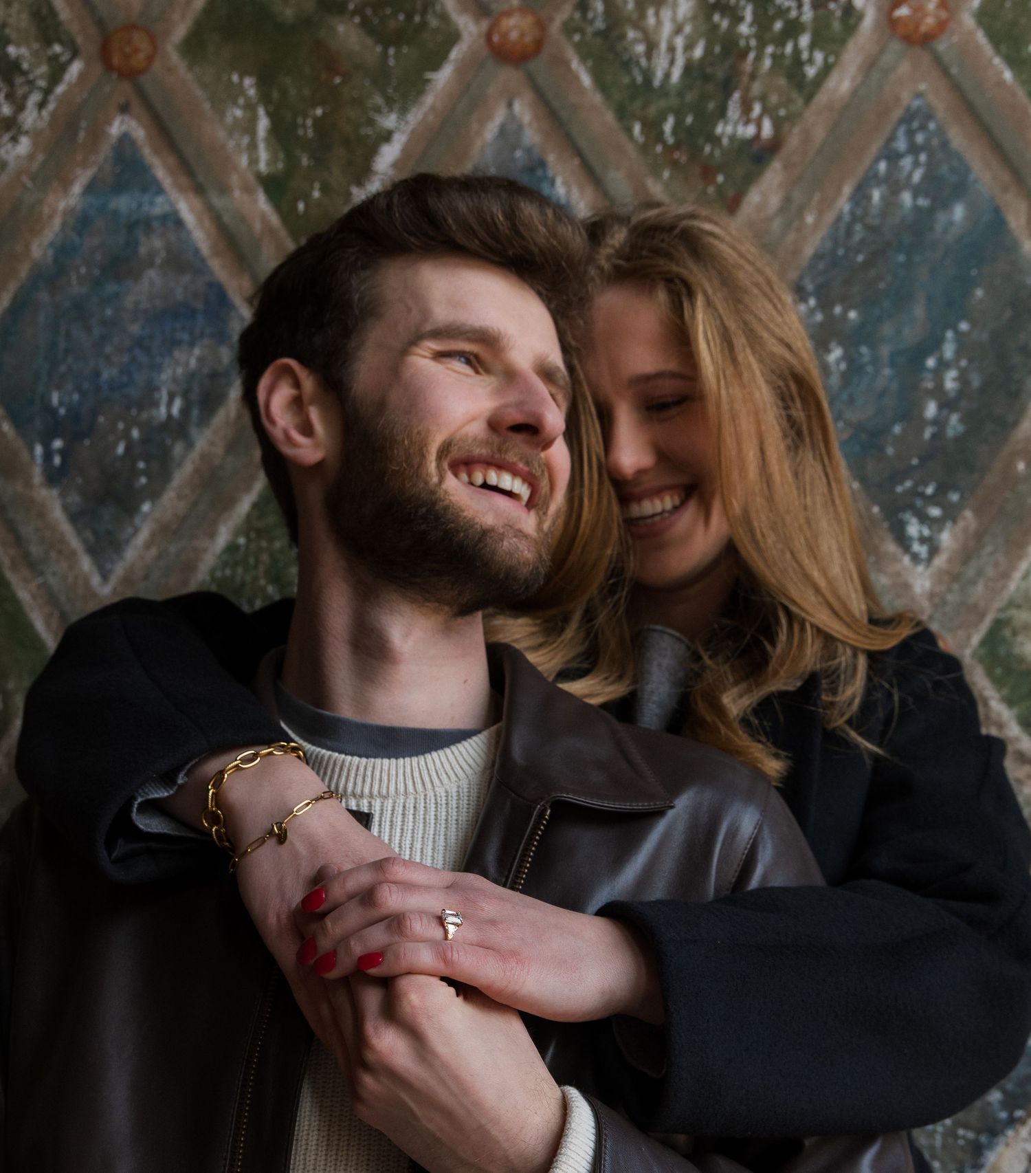 Happy couple embracing and smiling, woman with red nails hugging man from behind against decorative wall.