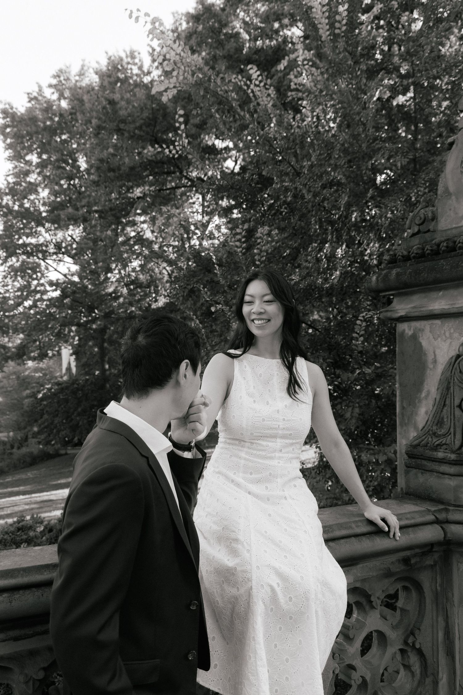 Black and white photo of a smiling bride in white dress sitting on stone balustrade with groom in suit at Central Park.