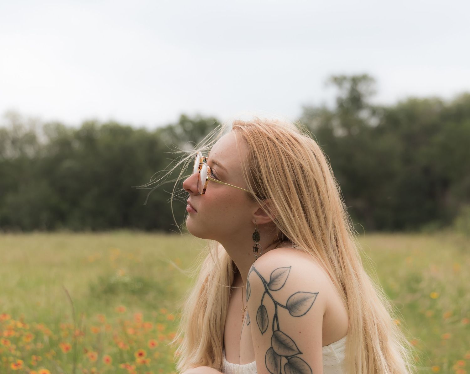 Side profile of blonde woman with leaf tattoo and glasses in wildflower field on cloudy day.