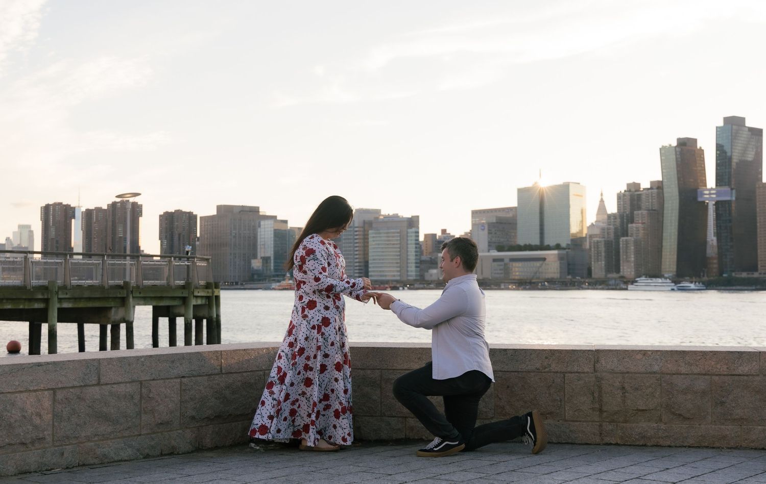Man proposes to woman on one knee by NYC waterfront skyline at sunset, romantic engagement moment.