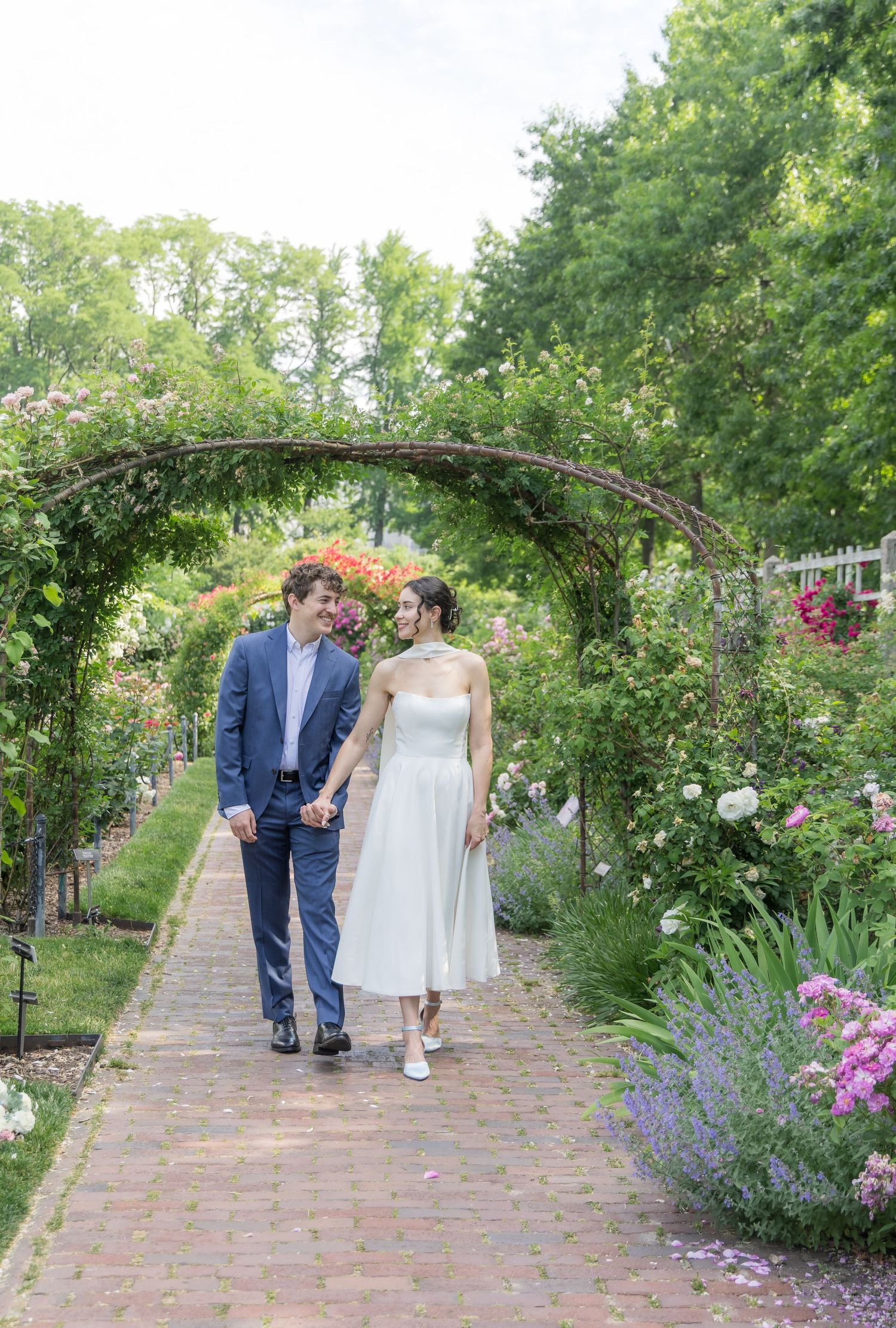 A couple walks hand-in-hand through a blooming garden archway, the woman in a white dress and the man in a blue suit.