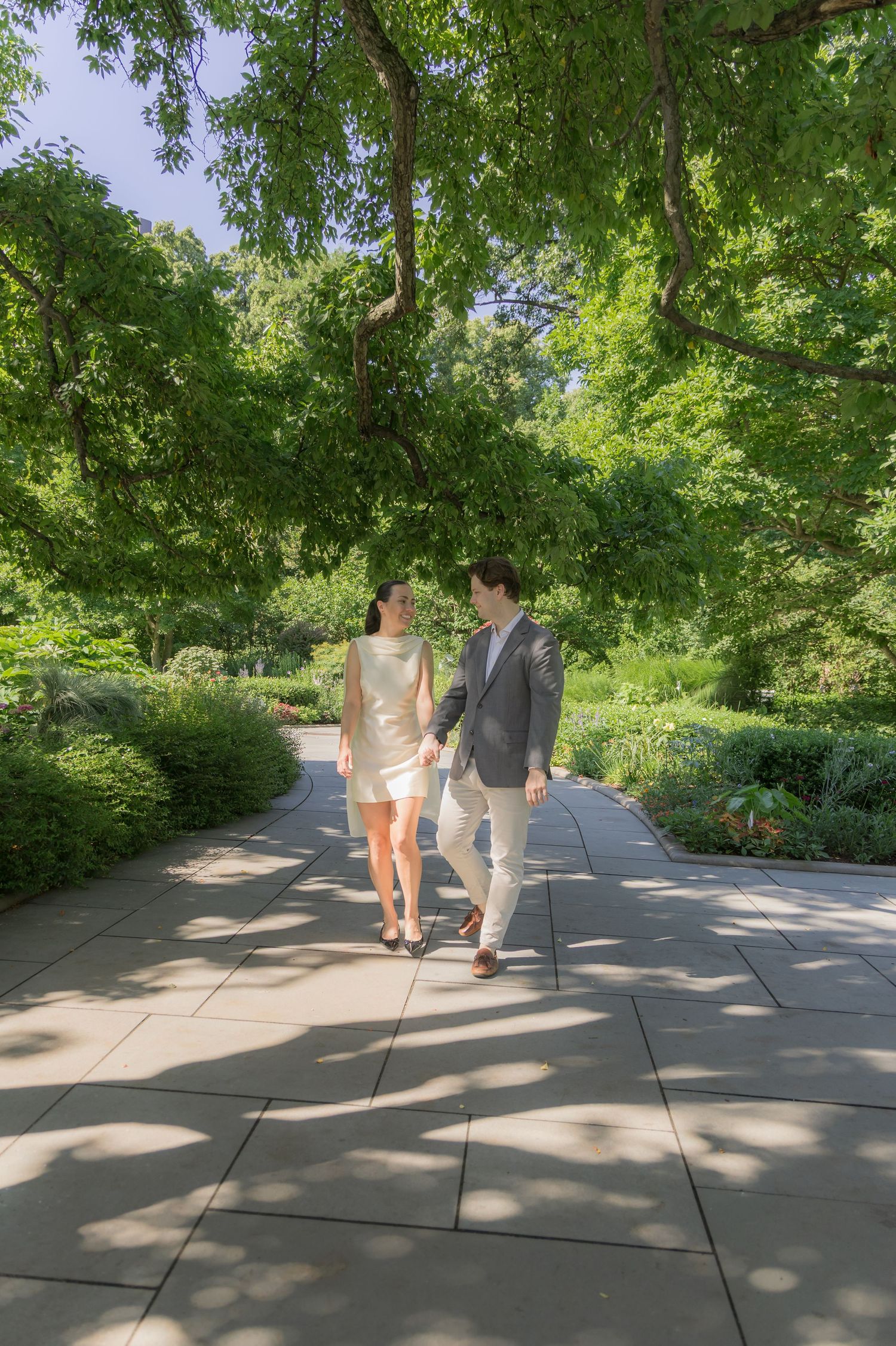 Couple walking hand-in-hand on a garden path surrounded by lush green trees and shrubs on a sunny day.